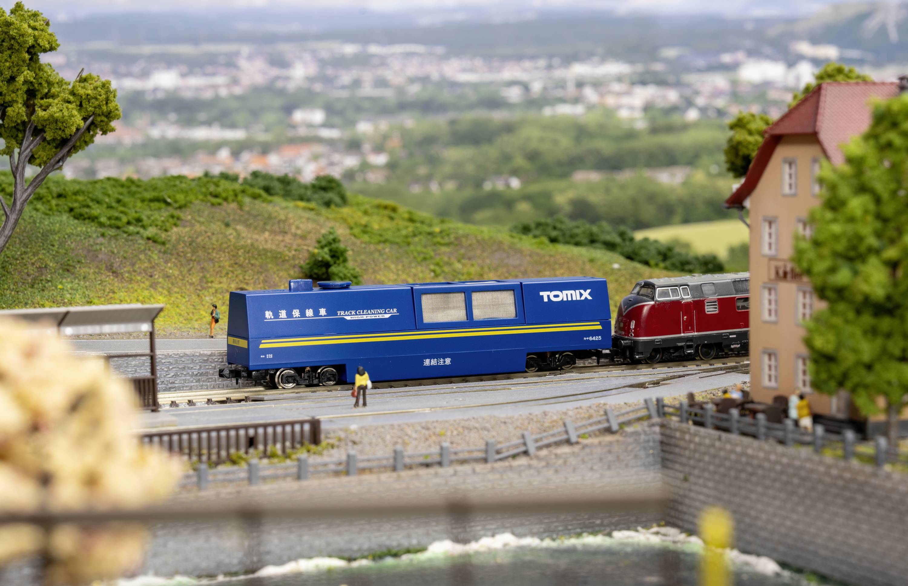 Model vláčku jede krajinou s zelenými kopci v pozadí. Modrý vůz 'Tomix' je viditelný.