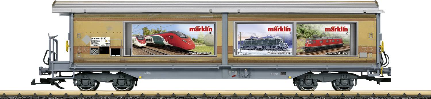 Model nákladního vozu s reklamou Märklin, zobrazující obrázky červených a modrých vlaků. Stojí na kolejích. Reklama na modelové železnice.