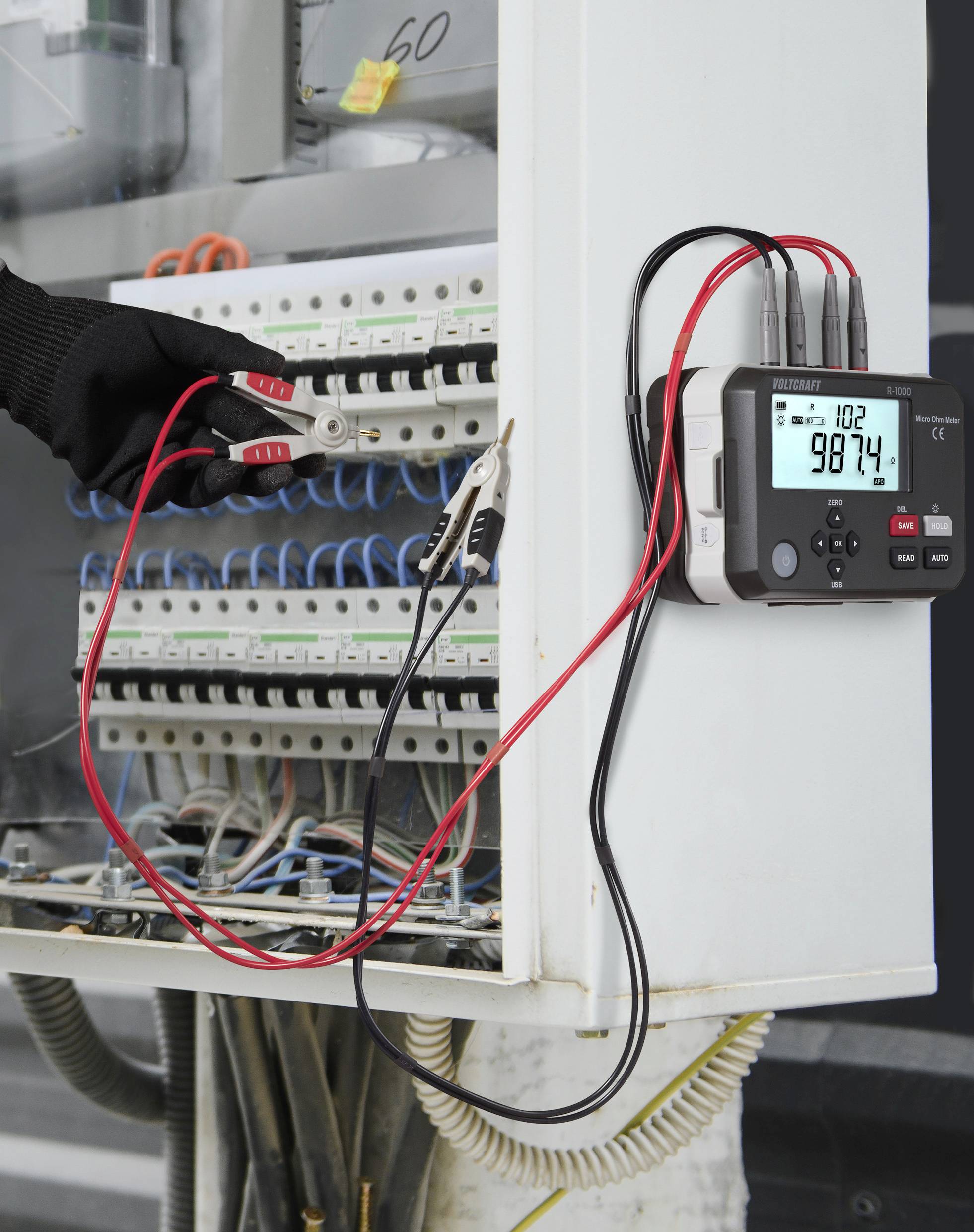Rukou inženýra v rukavici je pomocí červeného klešťového multimetru měřen proud v elektrickém rozvaděči, přičemž jeho digitální displej zobrazuje hodnotu '98,74'.