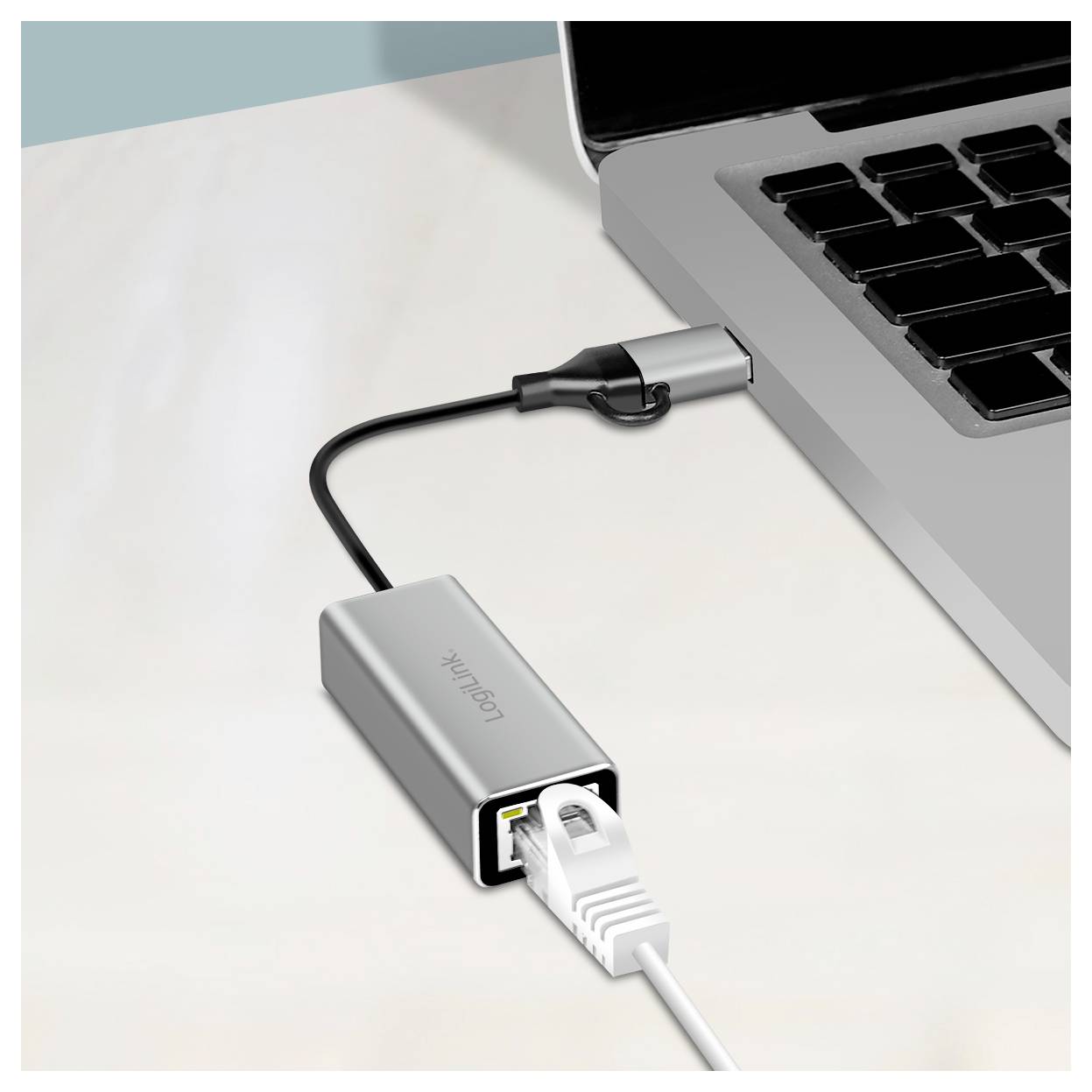 Laptop je připojen k síti prostřednictvím ethernetového kabelu a USB adaptéru. Adaptér je stříbrné barvy a kompaktní.
