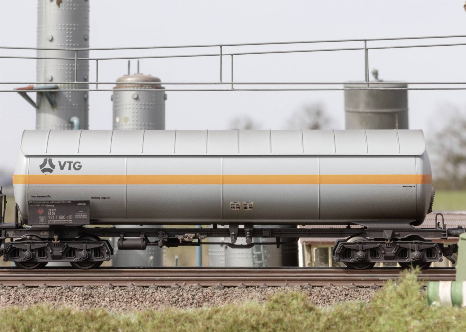 Stříbrný nákladní vagon s oranžovým pruhem a logem 'VTG' jede po kolejích, v pozadí jsou viditelné průmyslové struktury.