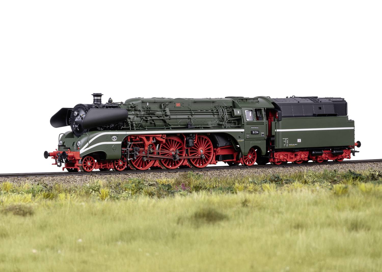 Zelený model parní lokomotivy s červenými koly na kolejích, obklopený trávou.