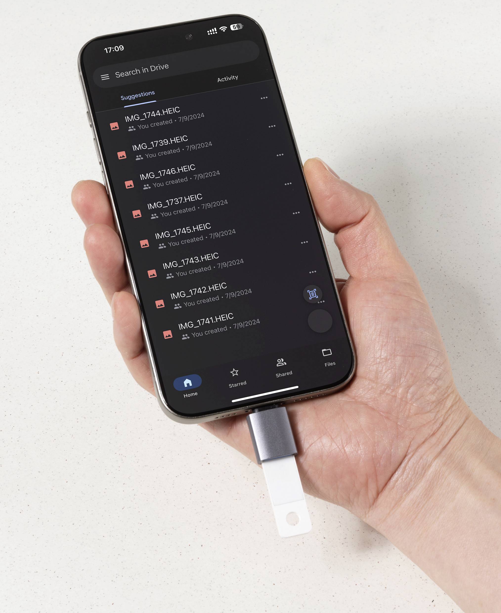 Ruka držící smartphone zobrazující seznam obrázkových souborů v cloudové úložné aplikaci. Připojený flash disk je připojen k telefonu.