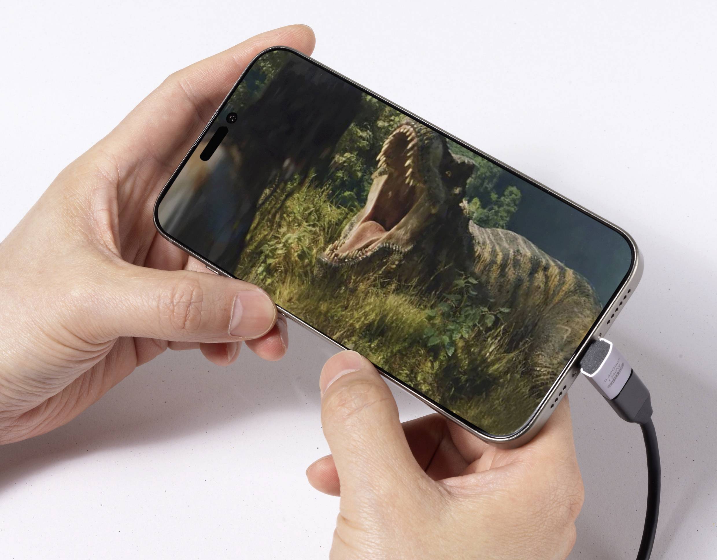 Osoba držící chytrý telefon zobrazující scénu řvoucího dinosaura v husté listnaté vegetaci, přičemž je telefon připojen k nabíjecímu kabelu.