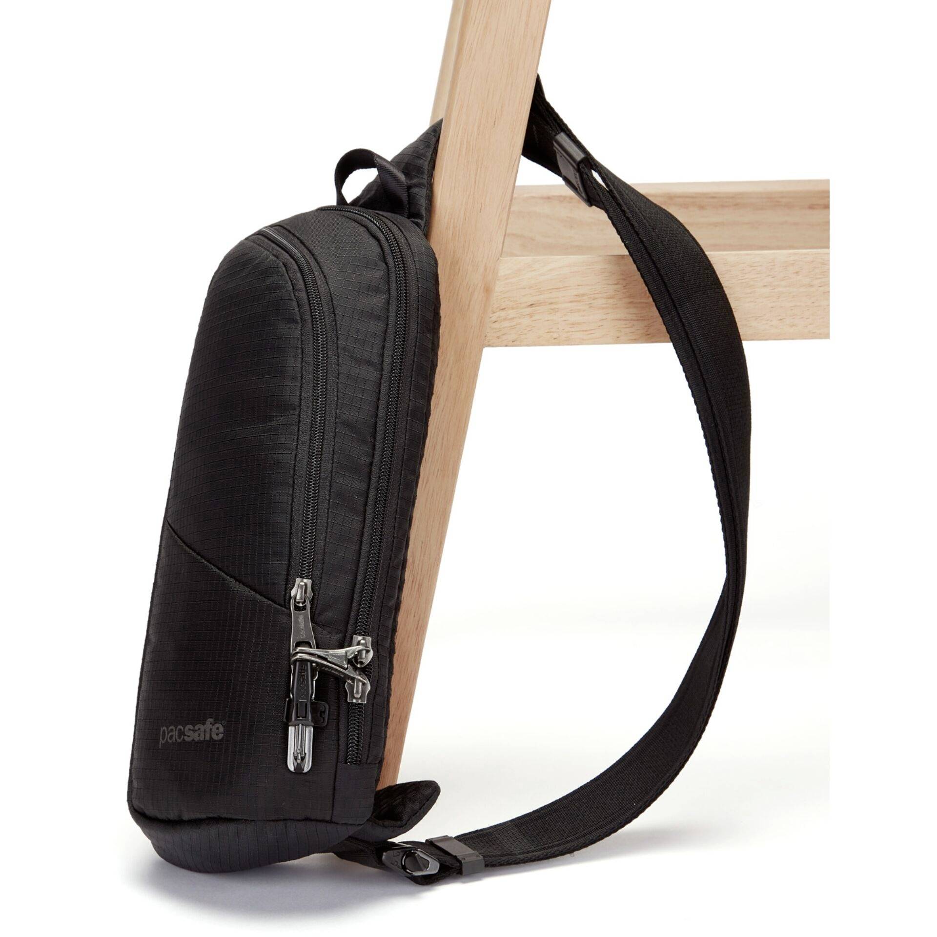 Černá crossbody taška je bezpečně připevněna k dřevěné noze stolu, což zdůrazňuje její protikrádežový design a bezpečnostní prvky.