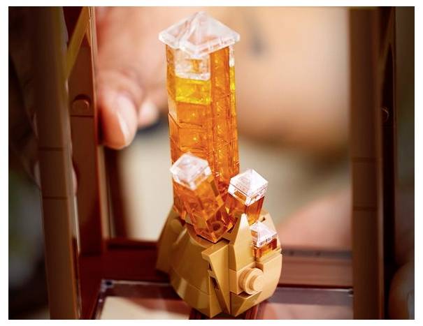 Detail záběru ruky držící Lego stavbu s třemi průsvitnými oranžovými krystaly na hnědé základně, symbolizující tvorbu drahokamů.