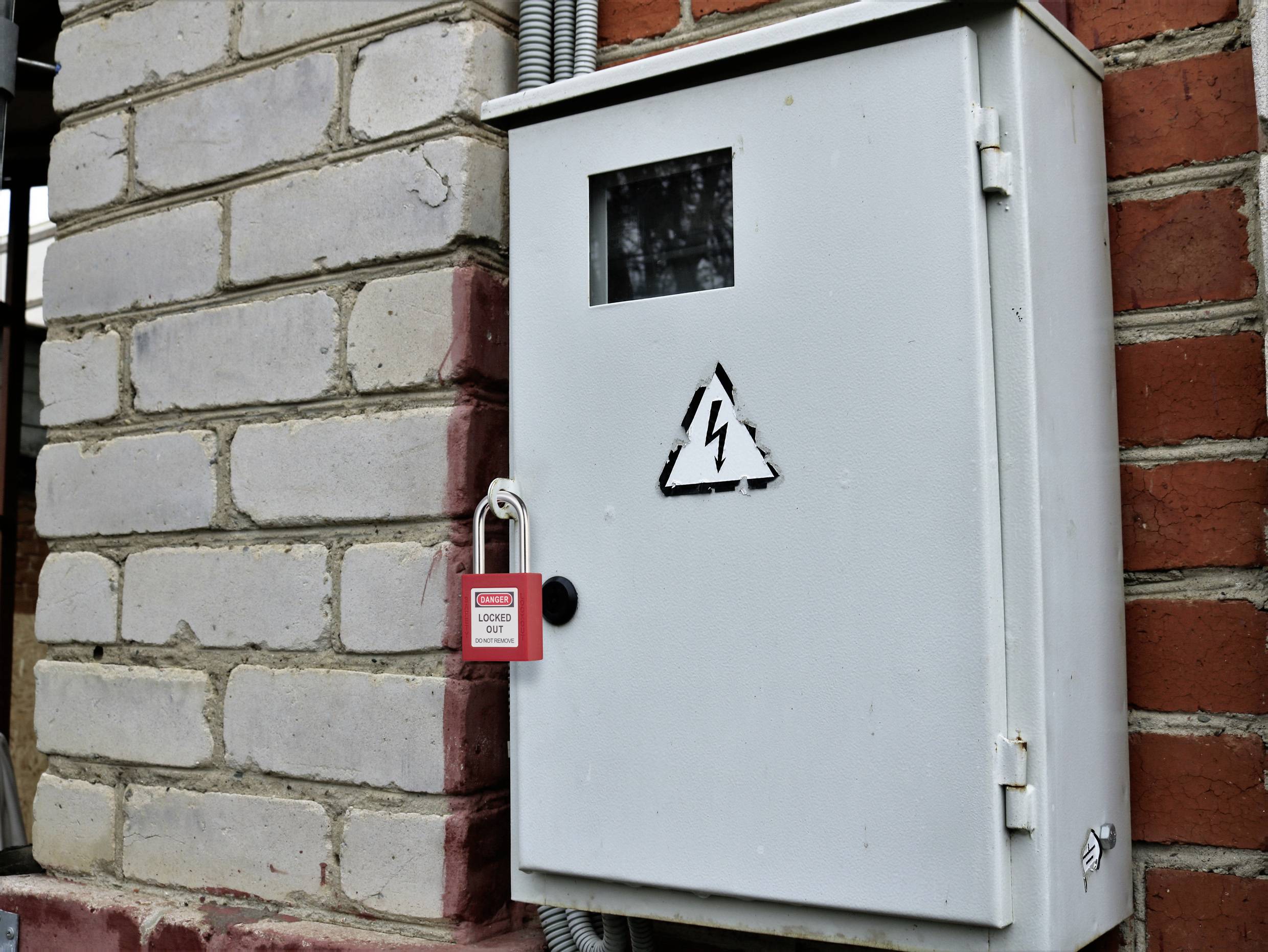 Elektrický rozvaděč s výstražným symbolem "Nebezpečí" uzamčený červeným visacím zámkem. Namontovaný na cihlové zdi, což signalizuje omezený přístup z bezpečnostních důvodů.