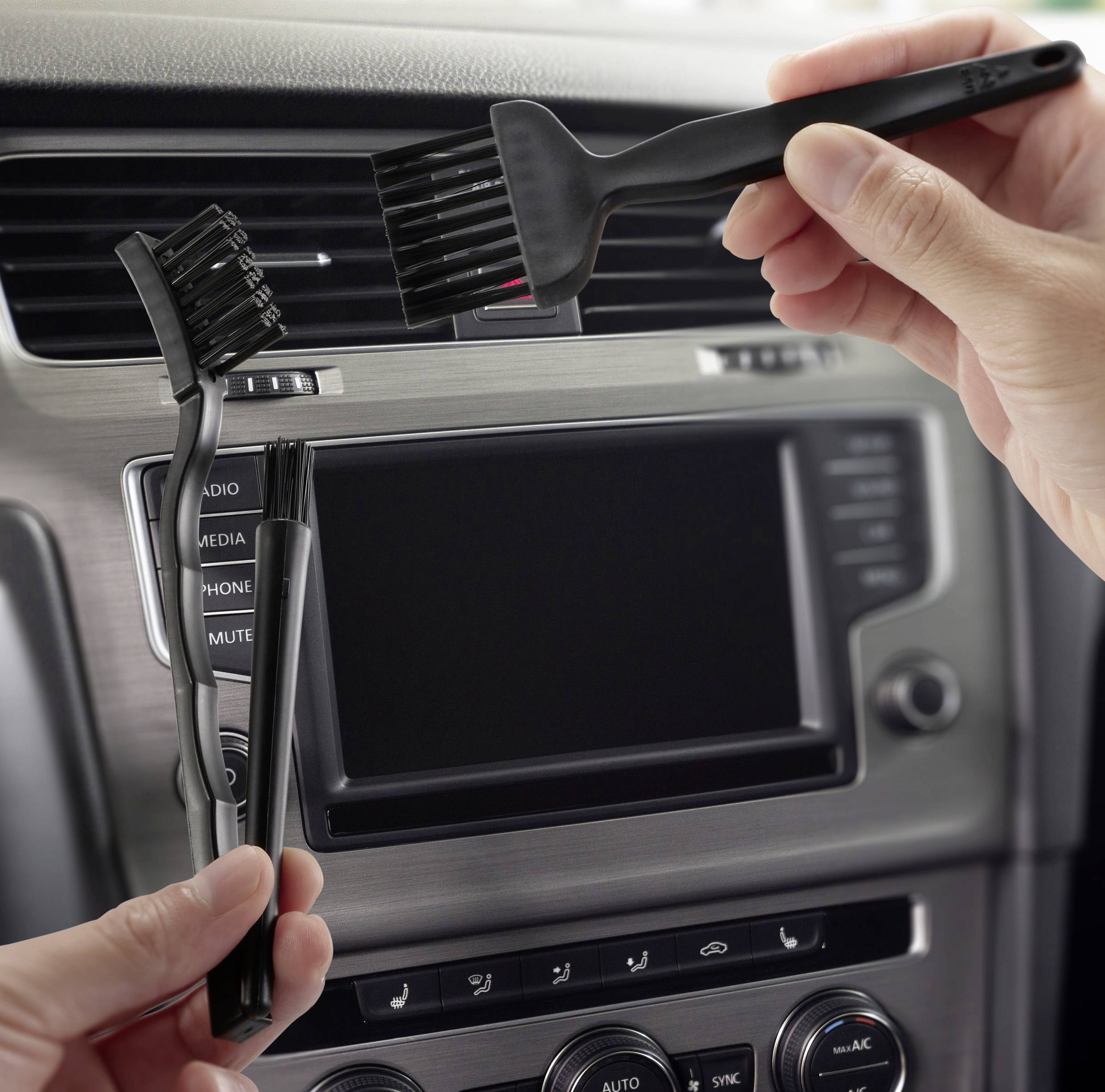 Ruce používající malý kartáček k čištění ventilačních otvorů v automobilu, s ovládacími prvky palubní desky viditelnými v pozadí.