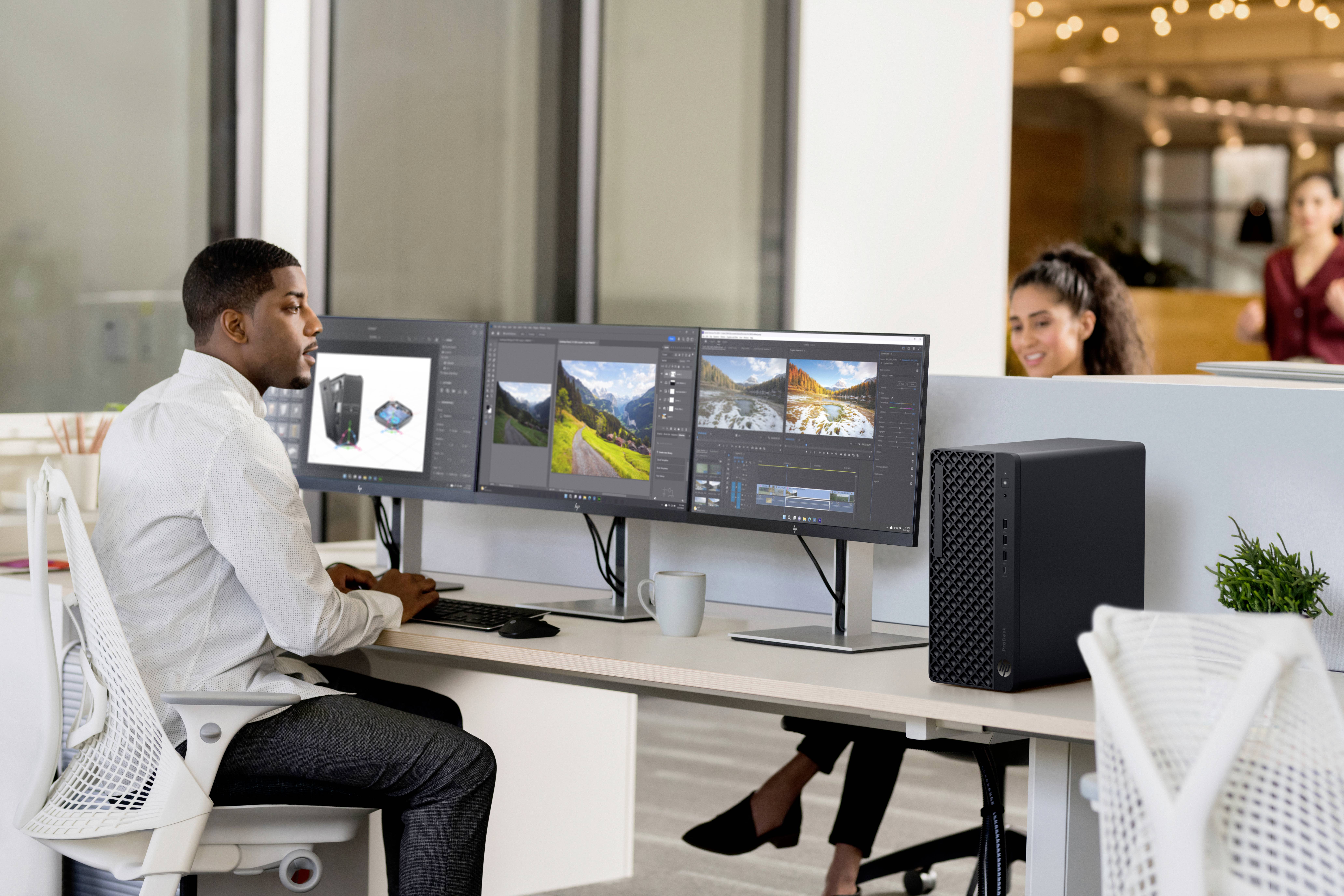 Osoba pracuje u stolu se třemi počítačovými monitory, na kterých je zobrazený designový software. V pozadní kancelářské oblasti je viditelná další osoba.