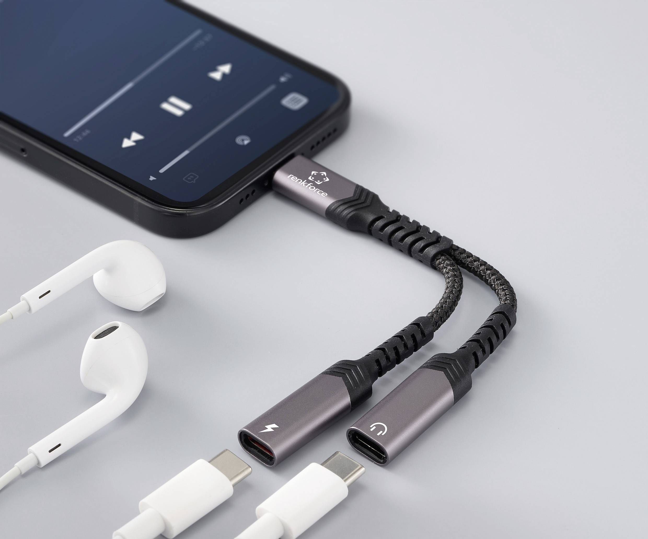 Chytrý telefon přehrávající hudbu je připojen k dvojitému adaptéru, který umožňuje současné použití sluchátek a nabíjení, vedle něj leží bílý nabíjecí kabel a sluchátka.