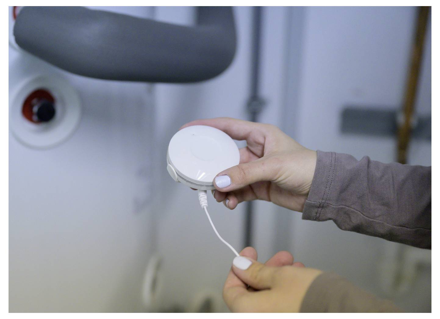 Ruce zapojující bílý nabíjecí kabel do malého kulatého zařízení vedle židle, pravděpodobně při přípravě lékařského nebo osobního vybavení.