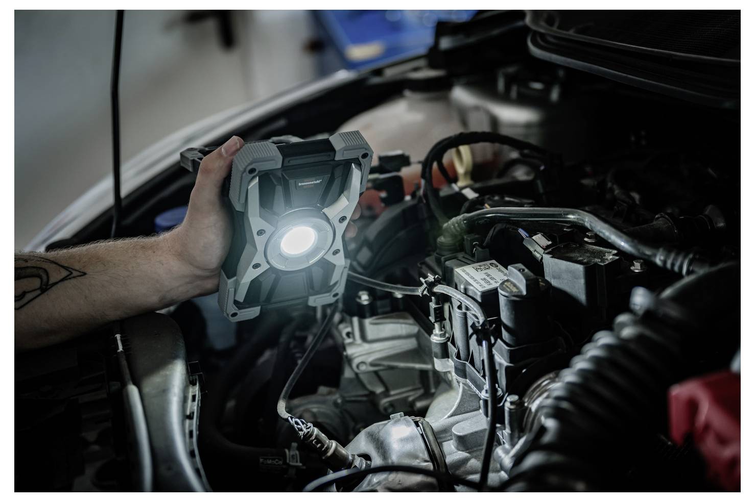 Osoba drží zapnutou pracovní lampu v motorovém prostoru automobilu, aby inspekovala nebo opravovala motor.