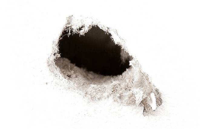 Malá, nepravidelná díra v bílém povrchu, s roztrhanými okraji, odhalující temnotu uvnitř.