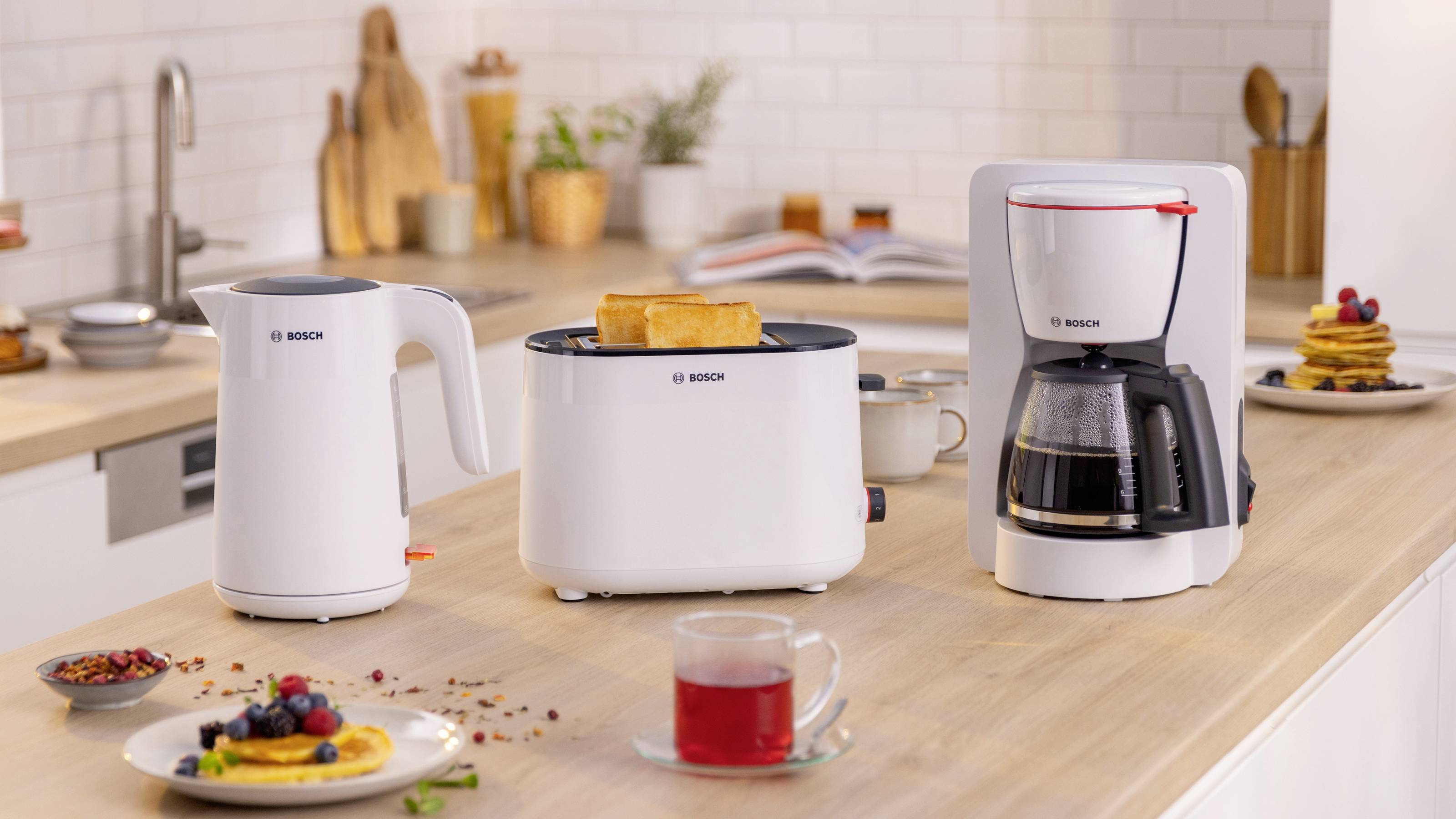 Moderní kuchyňská linka s bílou elektrickou konvicí, toustovačem s plátky toastů a kávovarem, obklopená snídaňovými pochutinami.