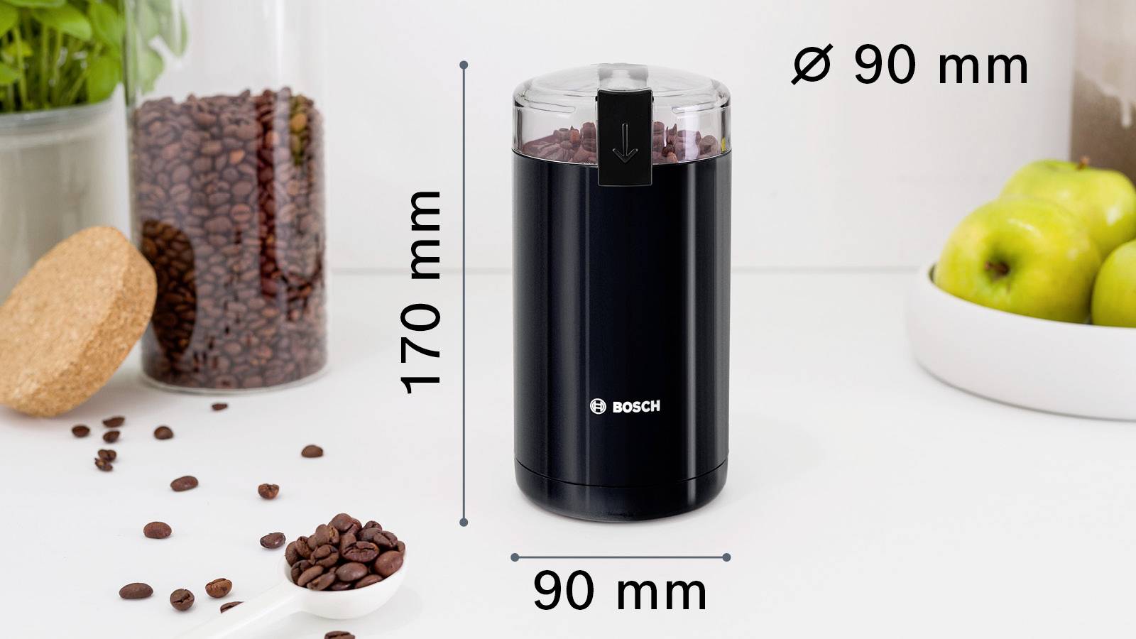 Černá mlýnek na kávu Bosch na kuchyňské lince s rozsypanými kávovými zrny, rozměry 170 mm výška a 90 mm průměr, vedle jablek.