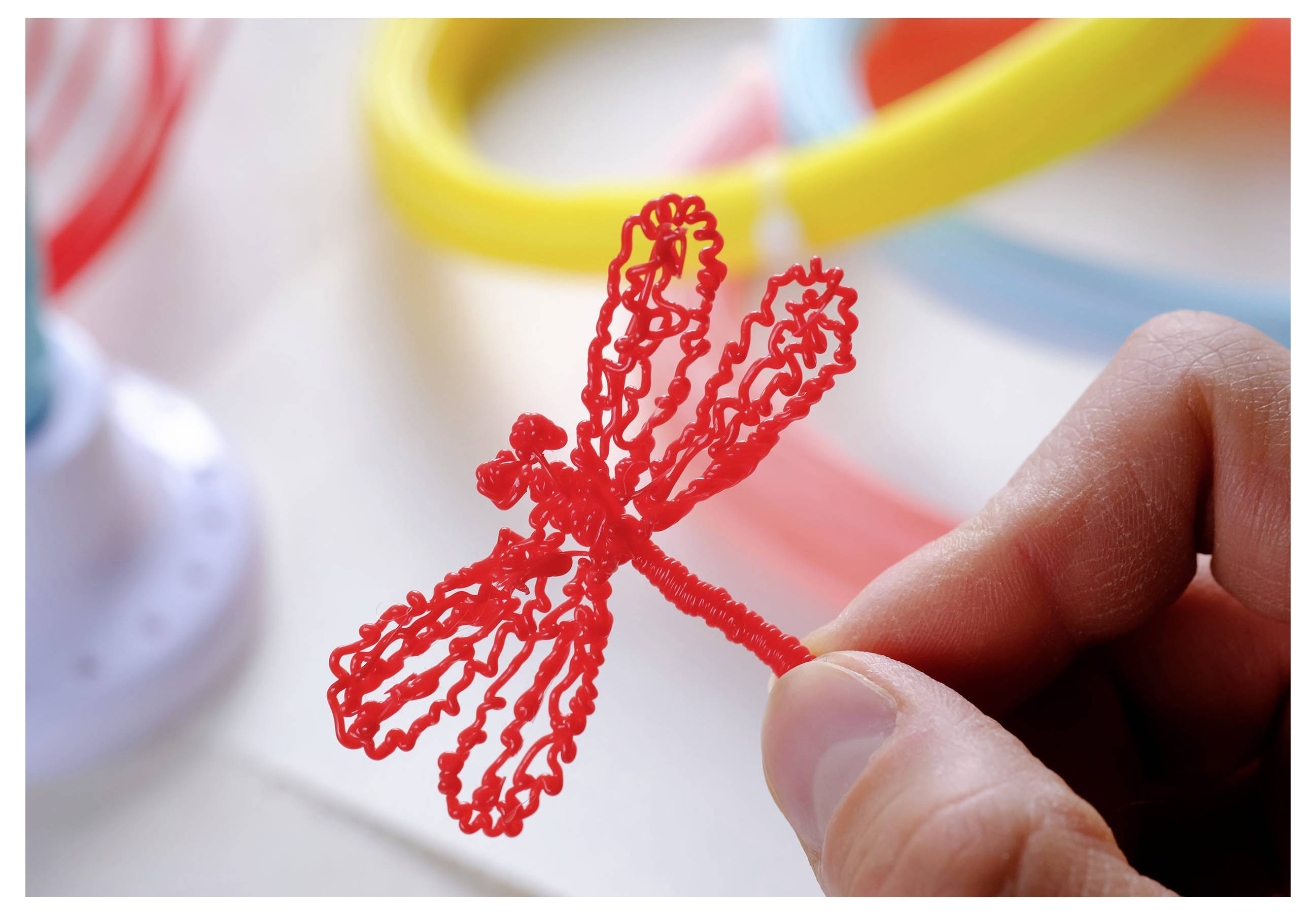 Ruka drží červený, precizně zkroucený plastový model vážky. V pozadí jsou barevné rozmazané trubice.