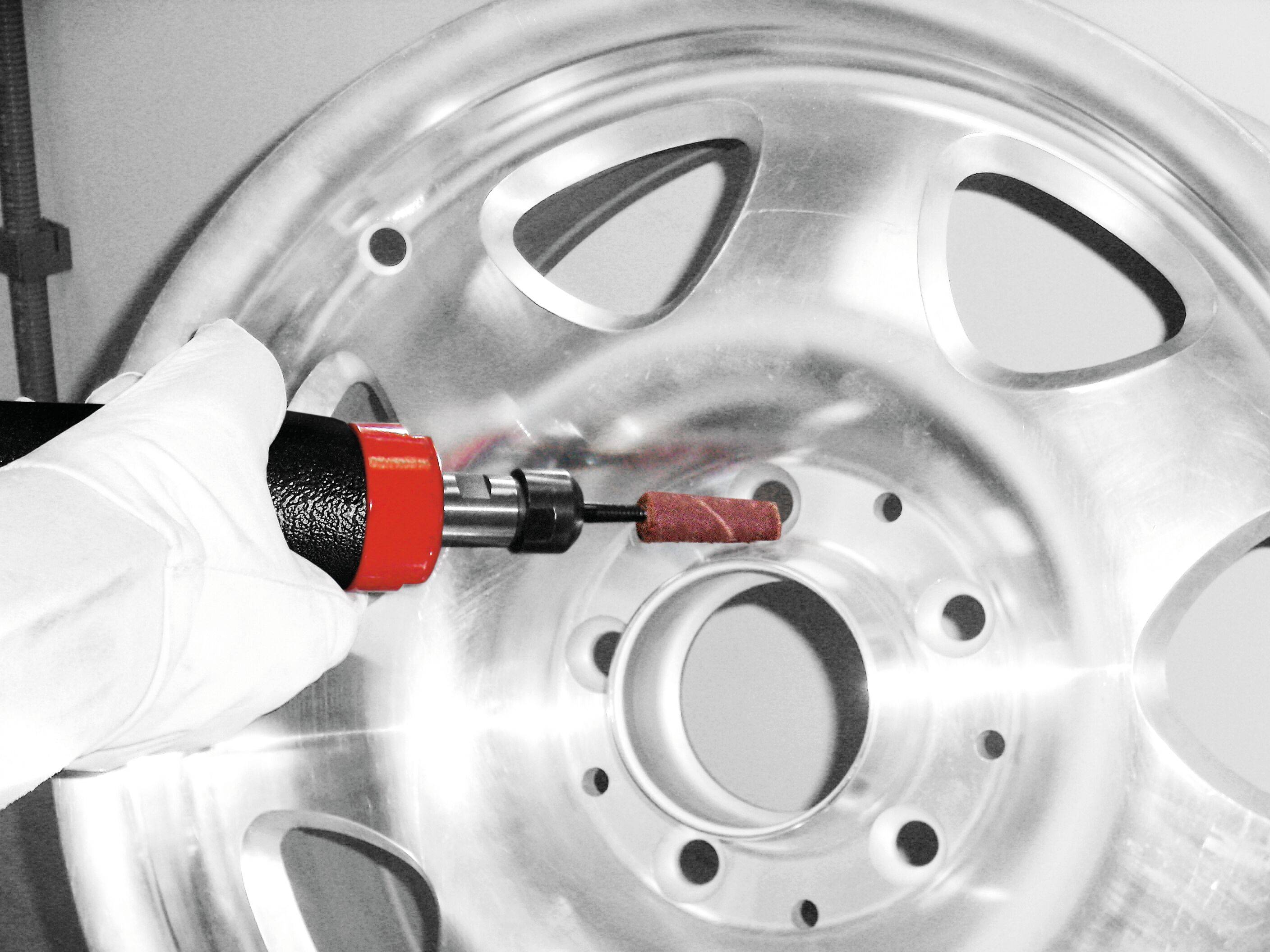 Rukavicí opatěná ruka používá červeno-černý elektrický nástroj k leštění lesklého stříbrného automobilového kola s několika výřezy.