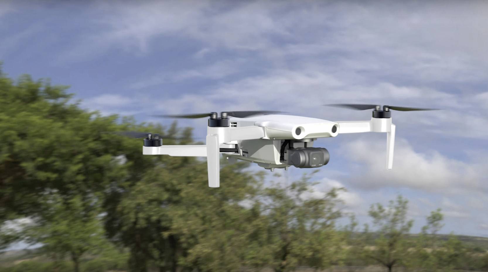 Bílý dron letící v parku s rozmazanými stromy a zatažnou oblohou v pozadí.