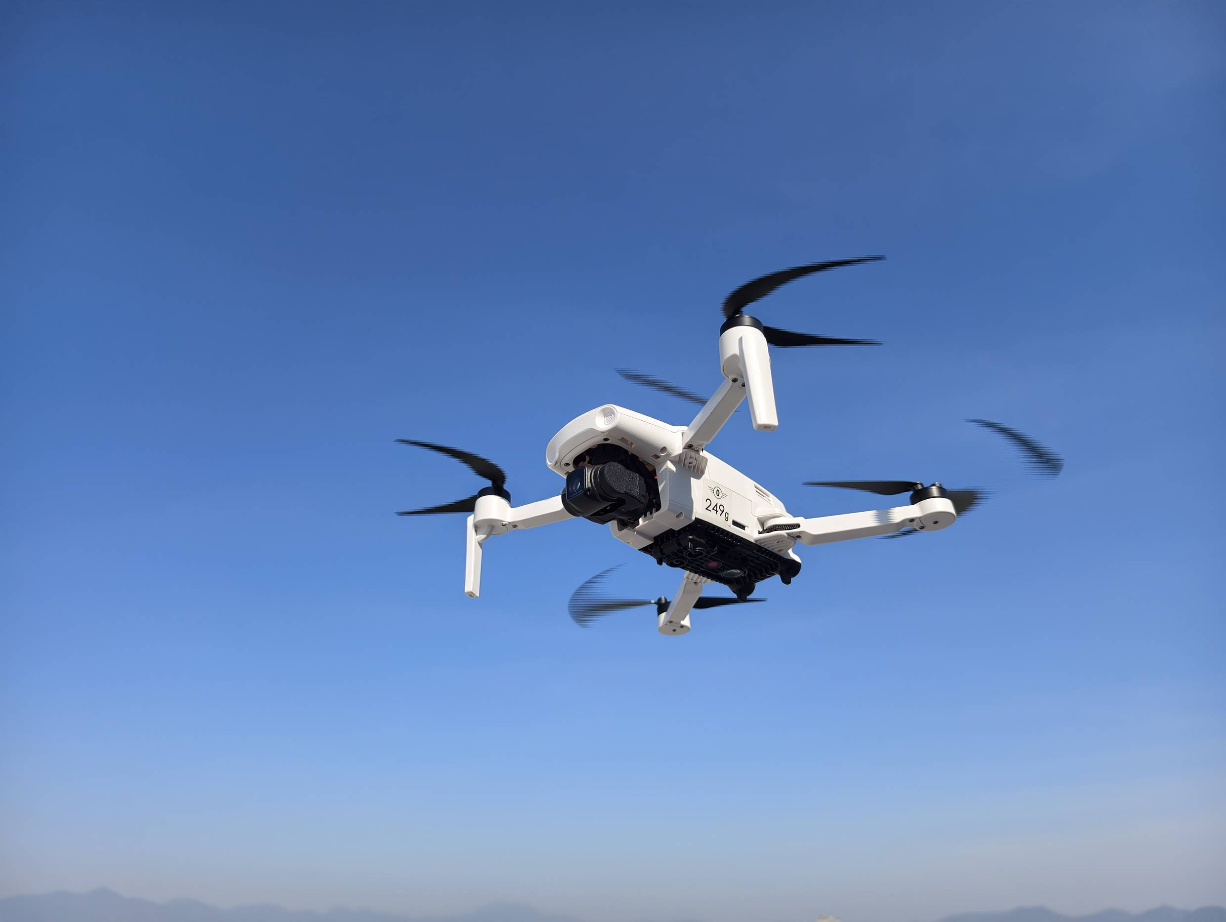 Bílý dron letící v čistě modré obloze, se dvěma viditelnými vrtulemi po stranách nakloněnými v diagonálním úhlu, naznačující pohyb vpřed.