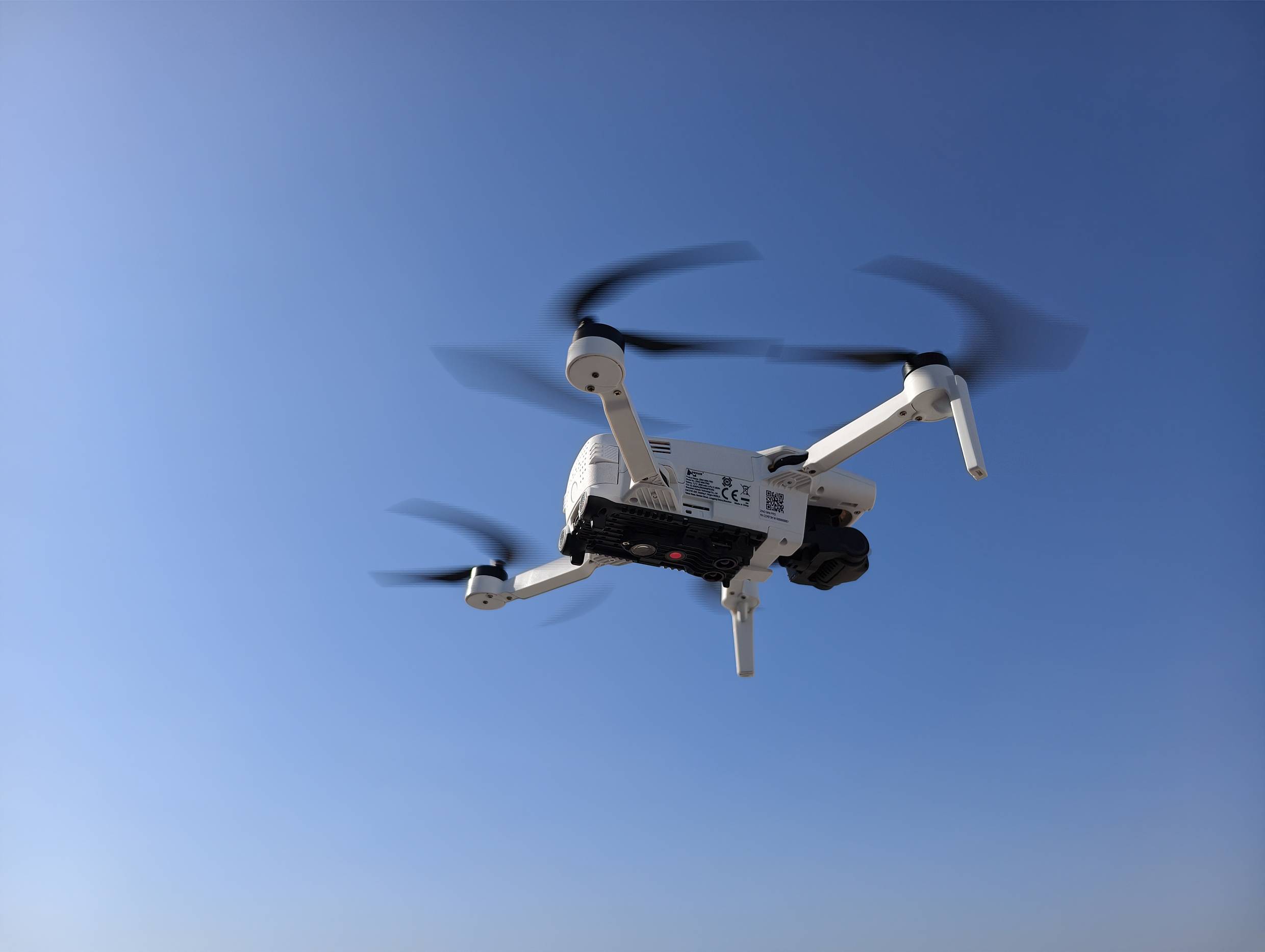 Bílý čtyřvrtulový dron letí v jasně modré obloze, jeho rotory rozmazané pohybem.