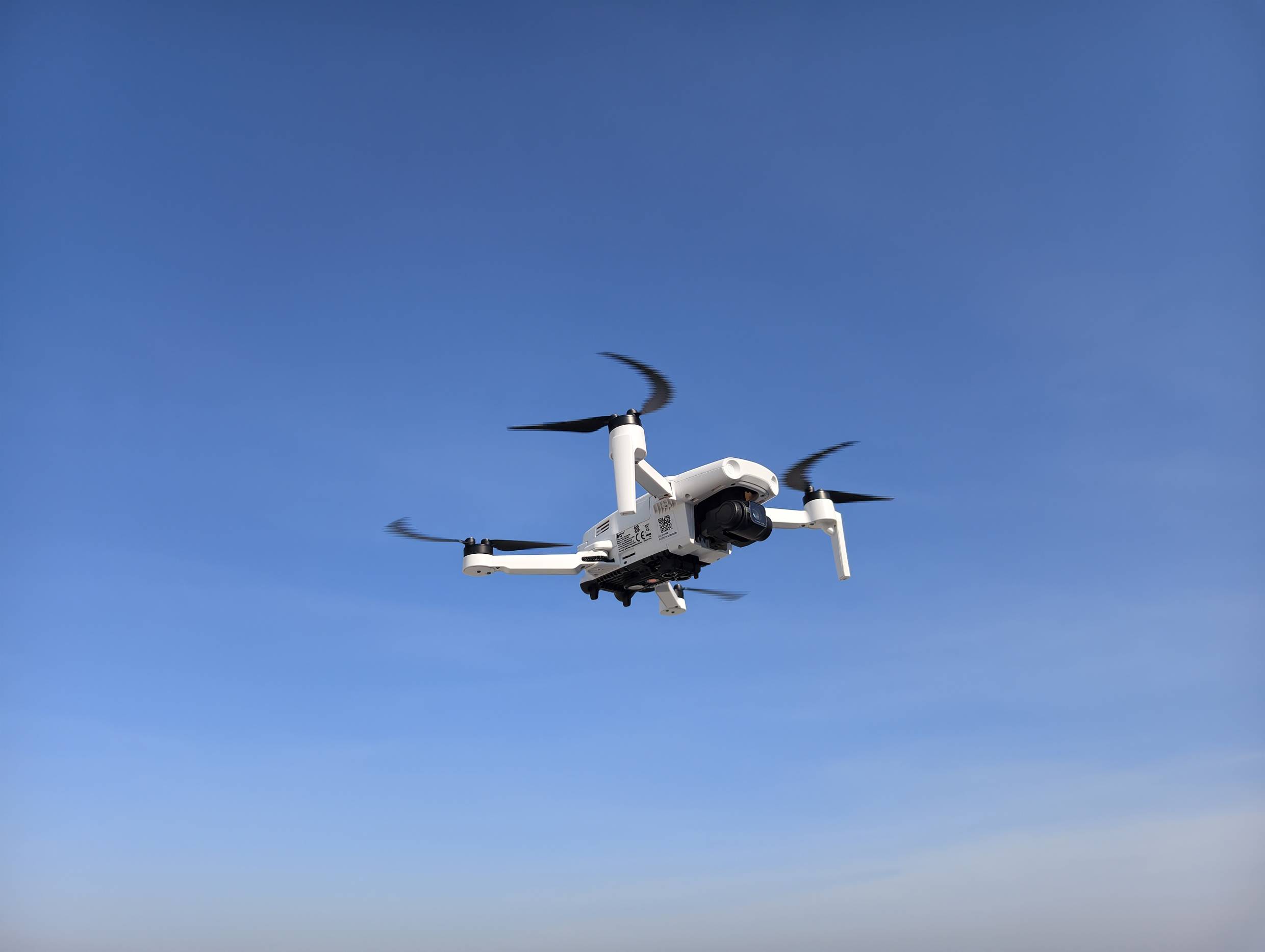 Bílý čtyřvrtulový dron letící proti jasně modré obloze, ukazující své vrtule a kameru, který naznačuje moderní leteckou technologii.
