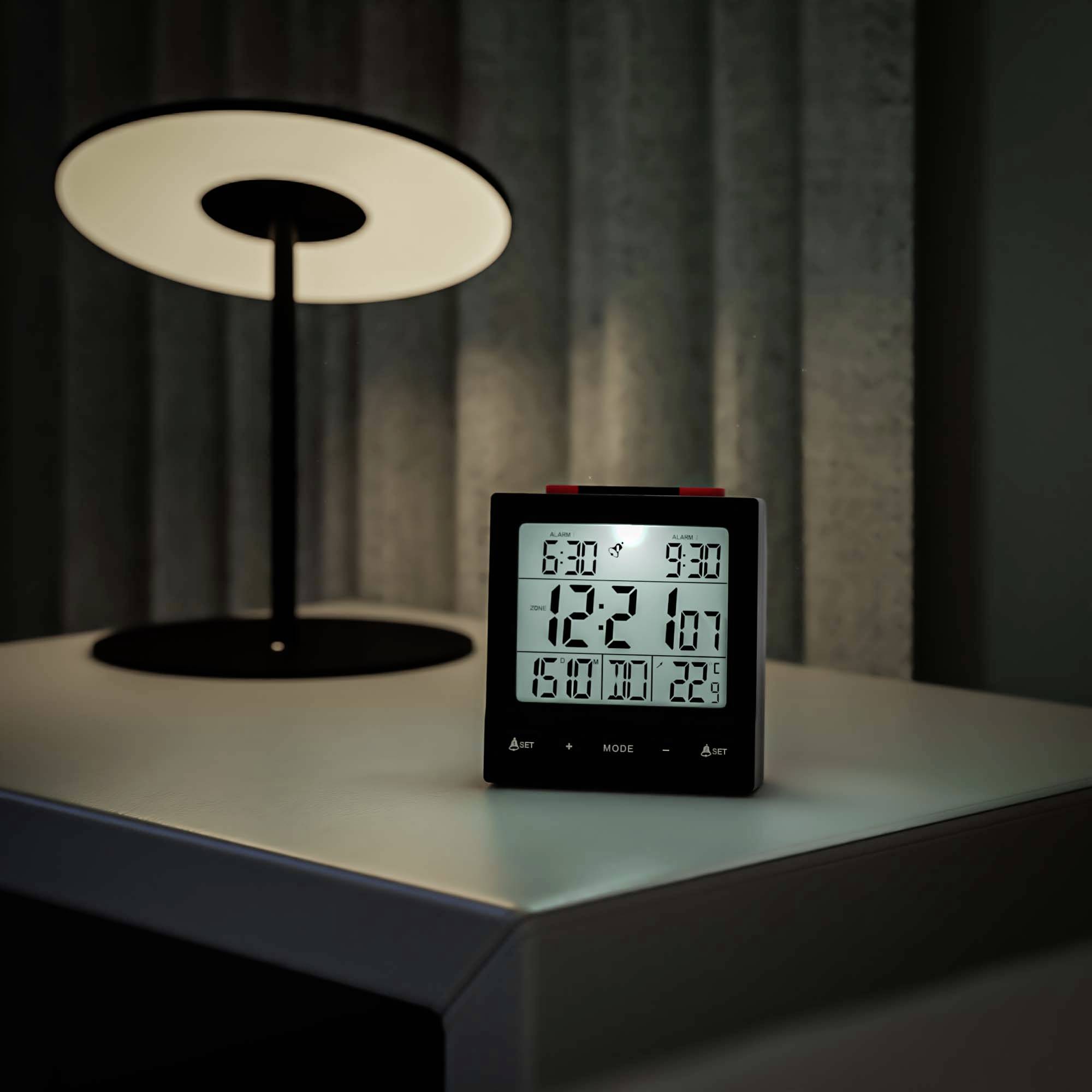 Digitální hodiny na nočním stolku ukazují '12:21', teplota je '29°C'. V pozadí svítí lampa s kulatým stínítkem.