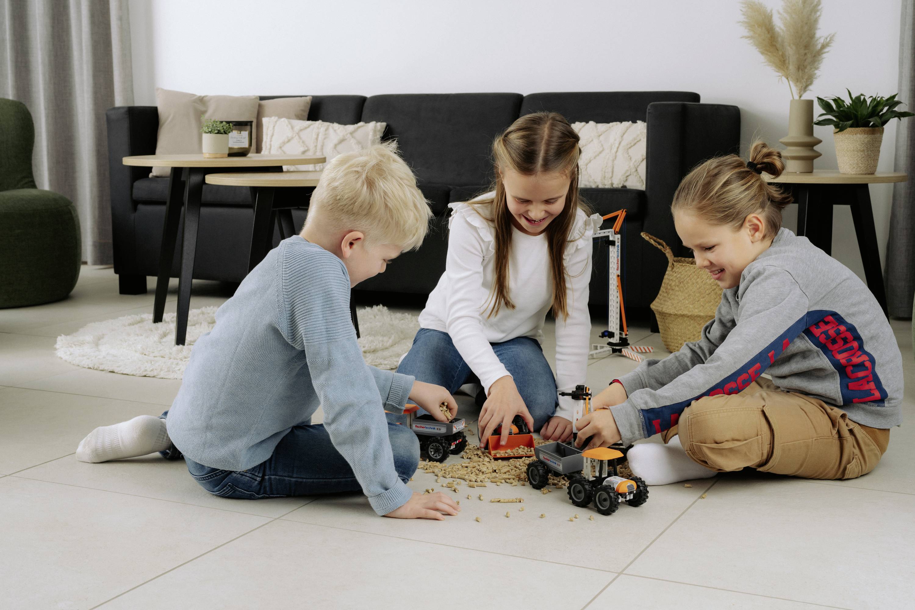 Tři děti si hrají s hračkářskými náklaďáky na podlaze v obývacím pokoji, usmívají se na sebe a vzájemně spolupracují, čímž vytvářejí roztomilou a hravou scénu.
