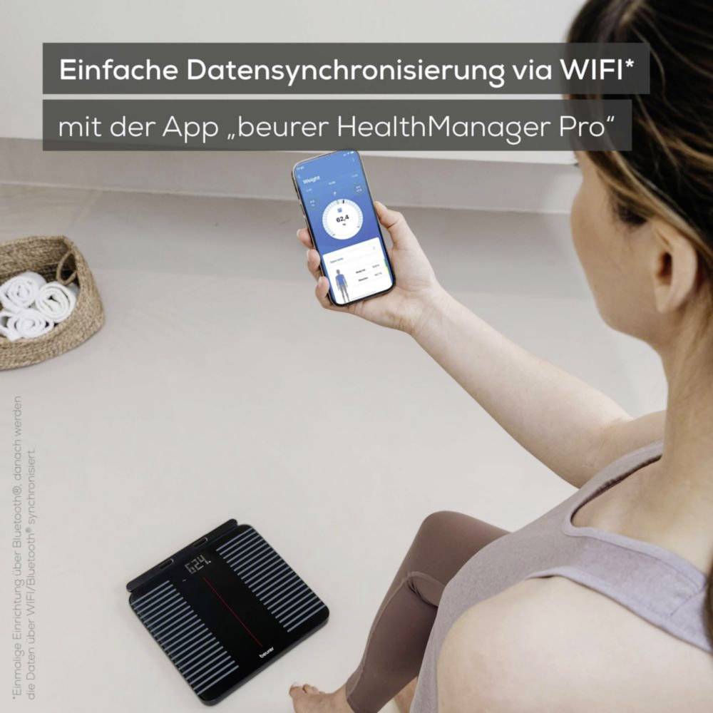 Osoba drží smartphone se zobrazenou aplikací 'beurer HealthManager Pro' pro synchronizaci dat pomocí WiFi. Vedle na podlaze stojí váha.