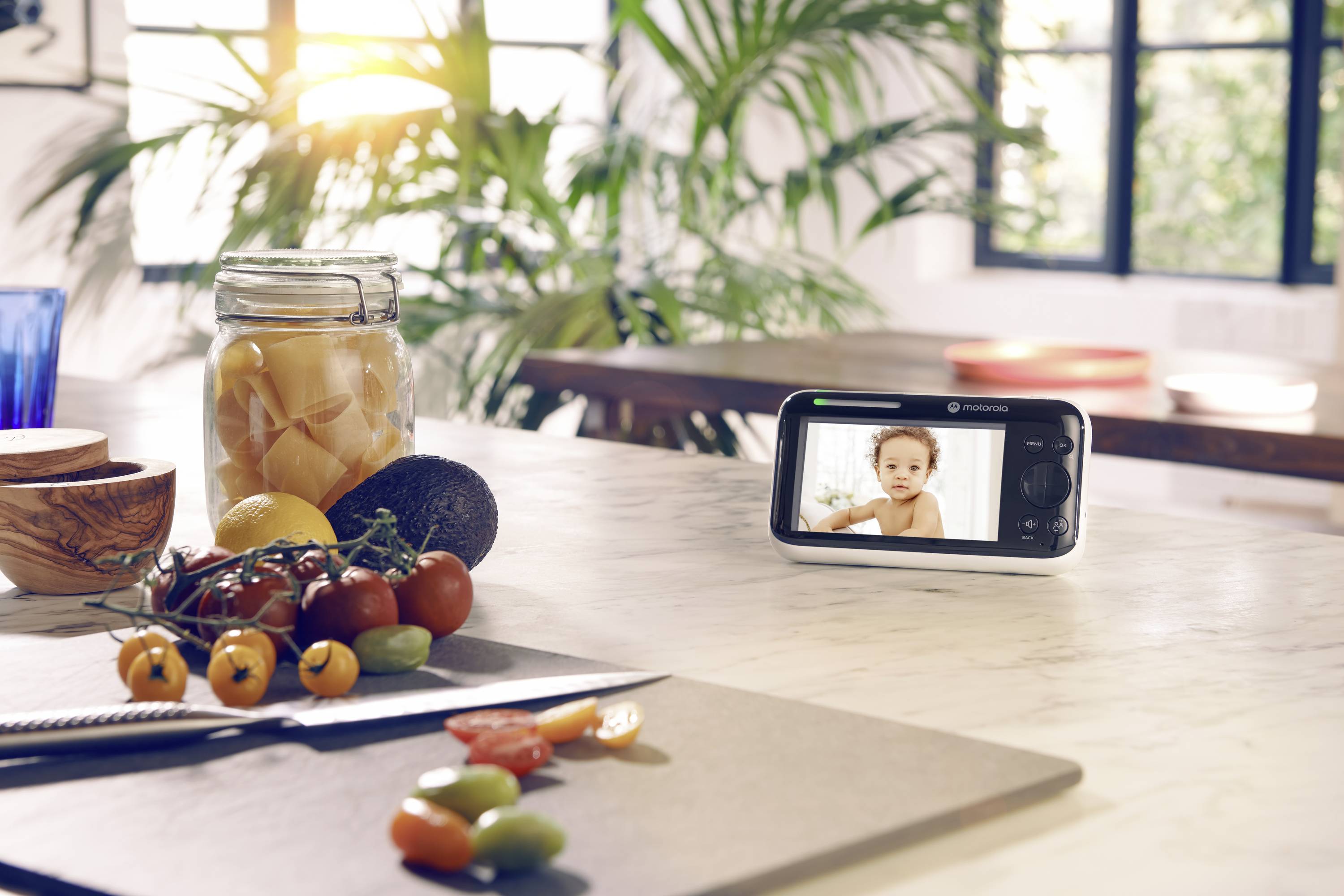 Kuchyňská linka s čerstvými cherry rajčaty, sklenicí zavařeniny a dětskou chůvičkou zobrazující dětský obraz. Sluneční světlo proniká dovnitř.