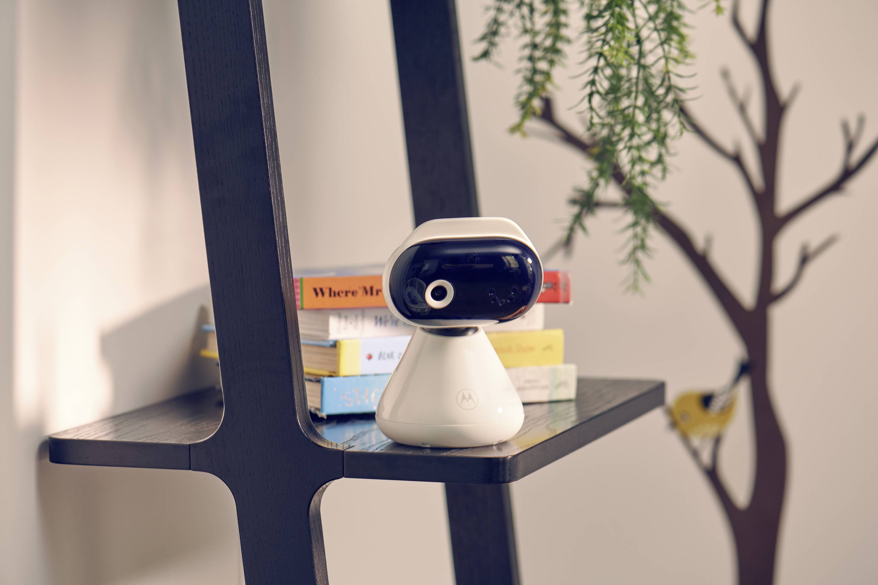 Malý, futuristický robotický fotoaparát stojí na dřevěné polici vedle naskládaných barevných knih, v pozadí je rostlina a nástěnná malba.