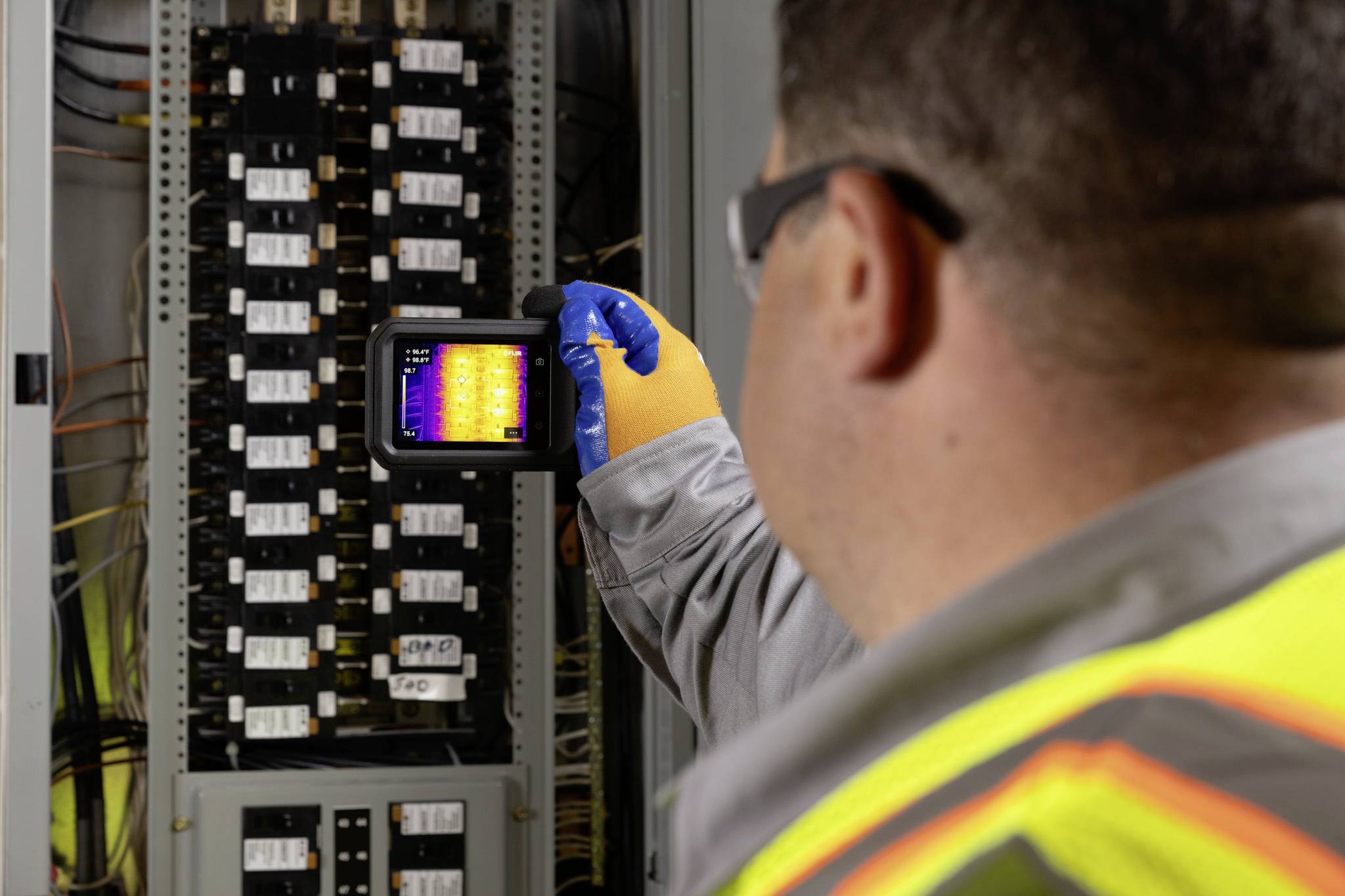 Technik v bezpečnostní vestě používá termální kameru k inspekci elektrického rozvaděče kvůli teplotním odchylkám.