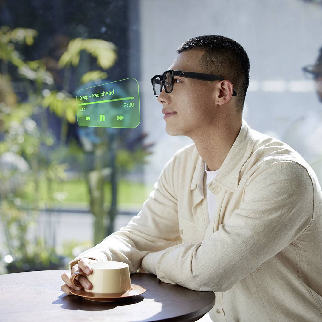 Muž v bílém saku sedí u stolu a pomocí rozšířené reality prohlíží rozhraní hudebního přehrávače, které zobrazuje 'Creep - Radiohead'.