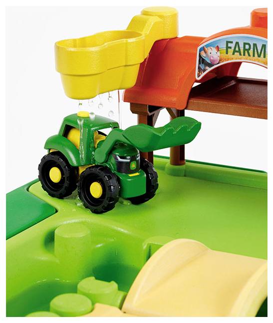 Malý zelený hračkářský traktor pod žlutou výpustí, na který stéká voda, vedle vstupu do hračkové farmy s cedulí.