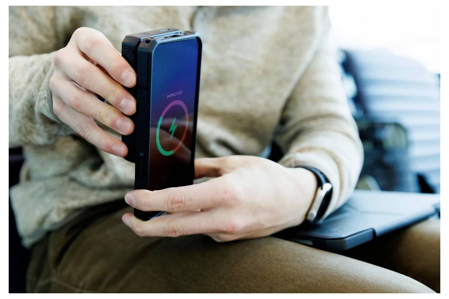 Osoba používající chytrý telefon s upozorněním na nízký stav baterie na obrazovce, sedící uvnitř, oblečená v béžovém svetru a hnědých kalhotách.