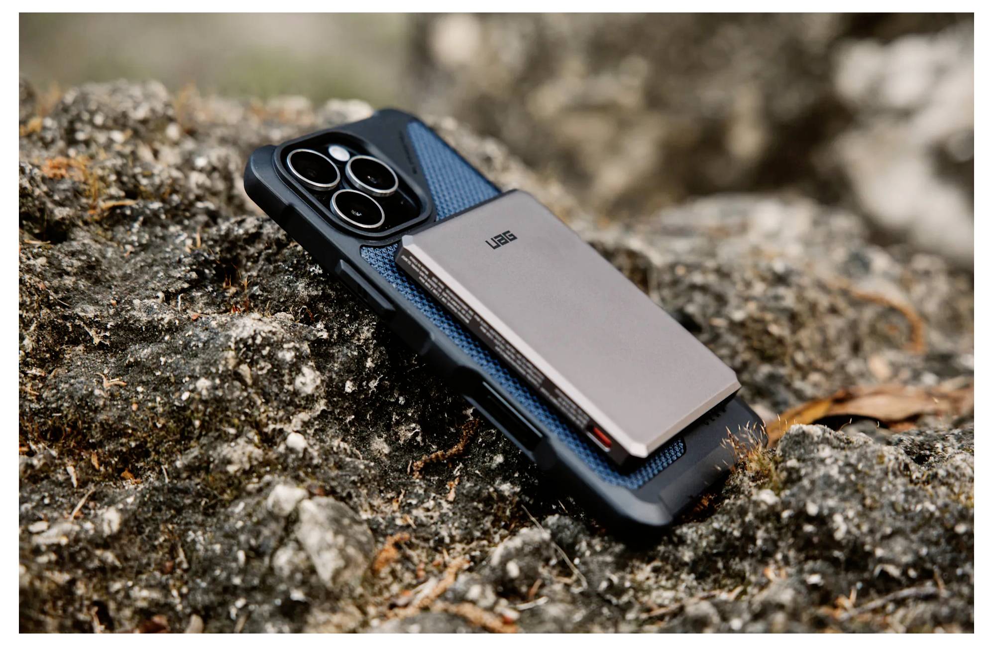 Chytrý telefon s ochranným pouzdrem a tenkým kovovým peněženkovým pouzdrem odpočívá na skalnatém povrchu venku.