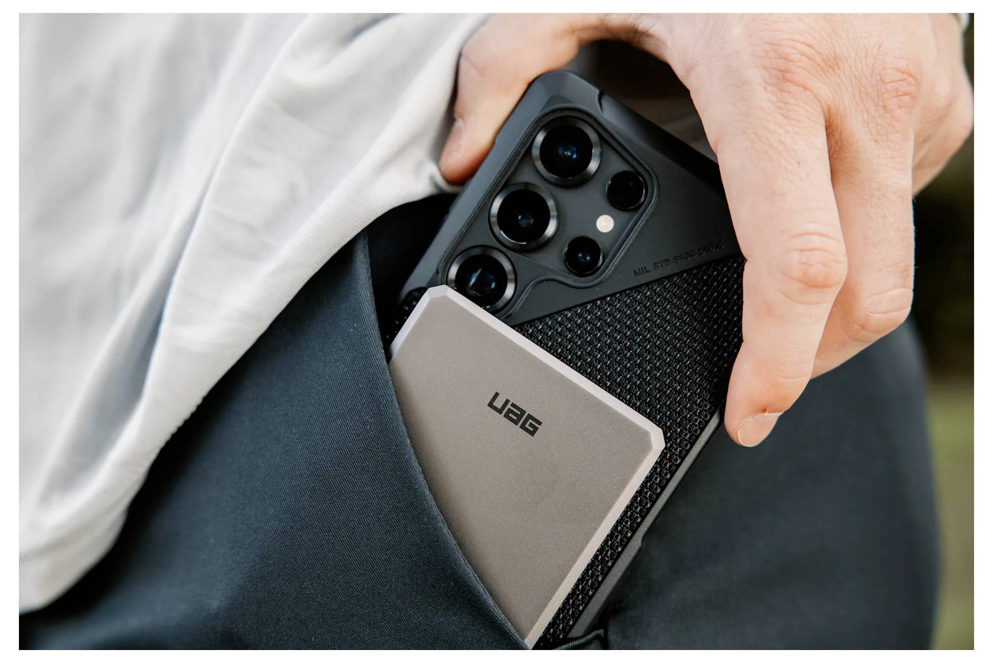 Osoba držící smartphone a zařízení označené jako "UAG" v kapse, s detailním pohledem na přenosné technologické přístroje.