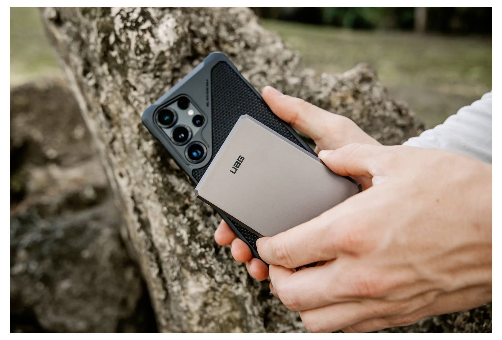 Osoba drží smartphone a přenosnou powerbанku u stromu v venkovním prostředí.