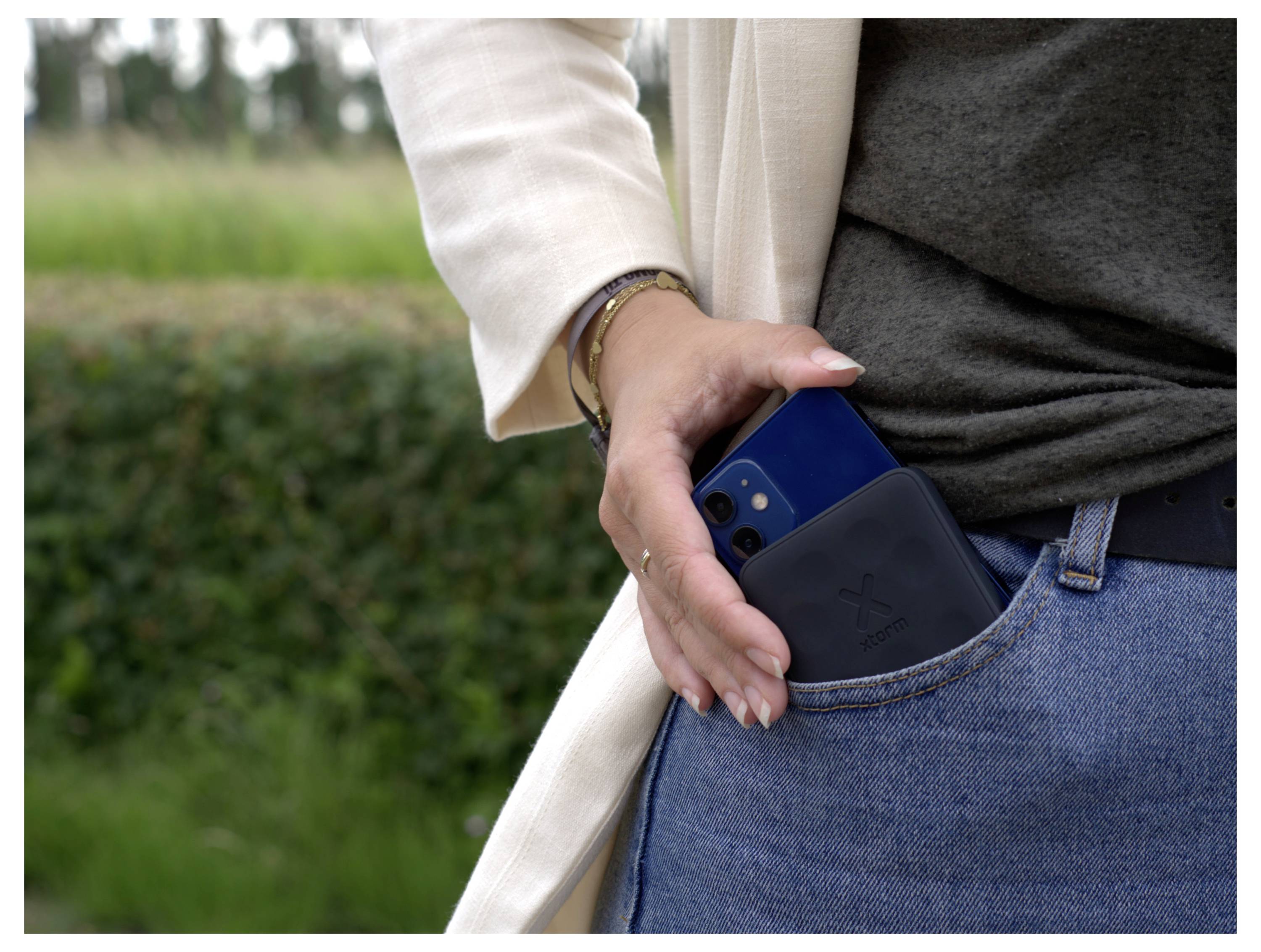 Osoba oblečená v bílé bundě a džínách vkládá smartphone do přední kapsy, na pozadí s rozmazaným venkovním prostorem.
