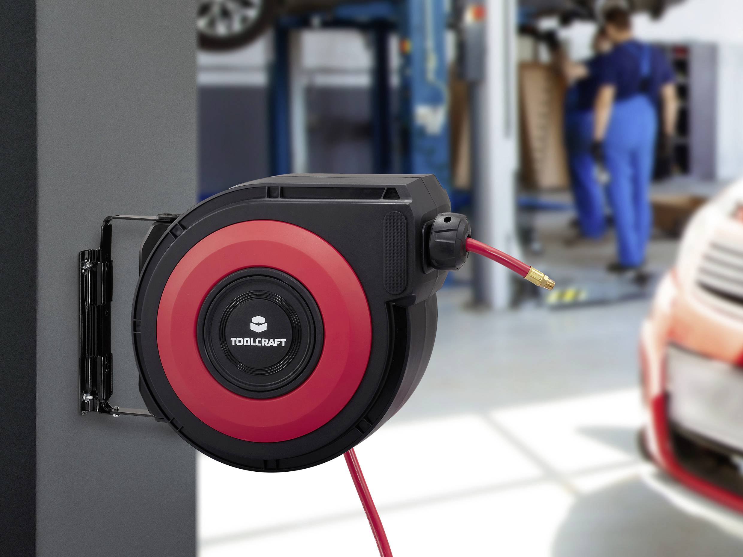 Červená a černá navíjecí hadice Toolcraft připevněná na stěně v automechanické dílně, v pozadí pracující mechanik.