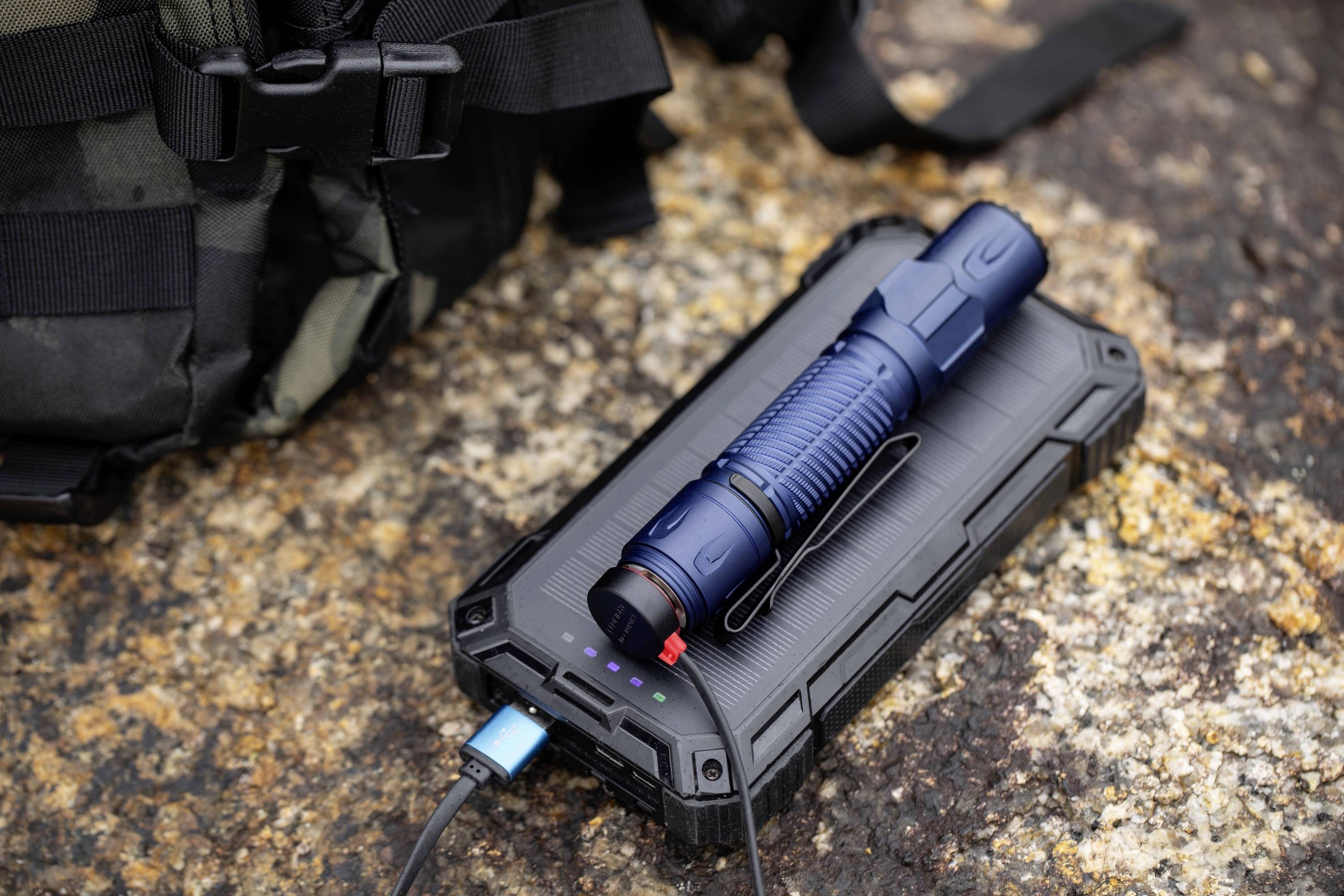 Modrá svítilna leží na robustní černé powerbance, propojené USB kabelem, umístěné na strukturovaném kameni vedle batohu.