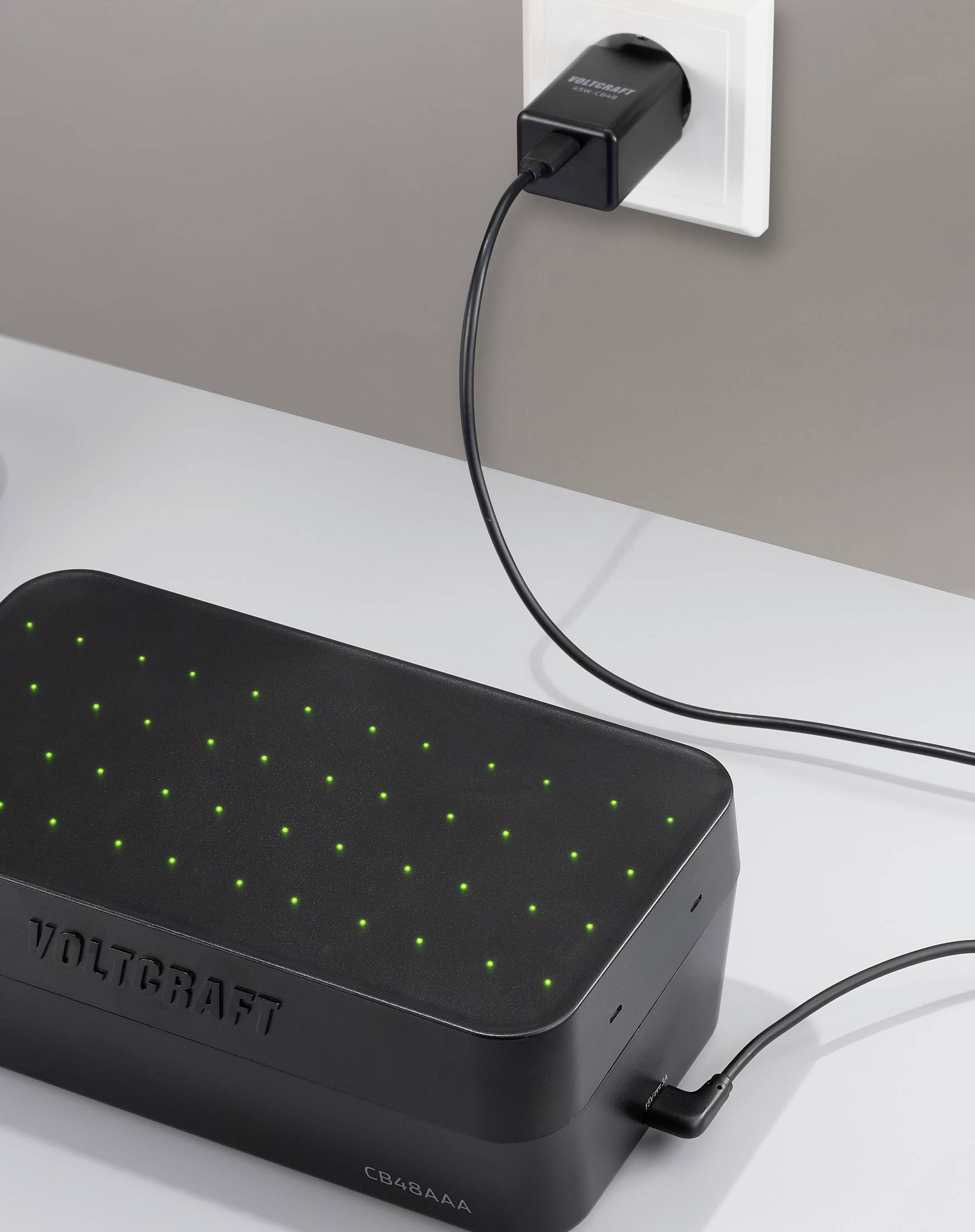 Černé obdélníkové zařízení se zelenými světly, označené štítkem 'Voltcraft', je zapojeno do elektrické zásuvky pomocí USB kabelu na šedém povrchu.