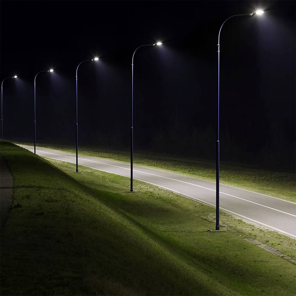 Řada pouličních lamp osvětluje křivolakou, prázdnou silnici v noci, obklopenou trávou a tmou, která navozuje pocit osamělosti.