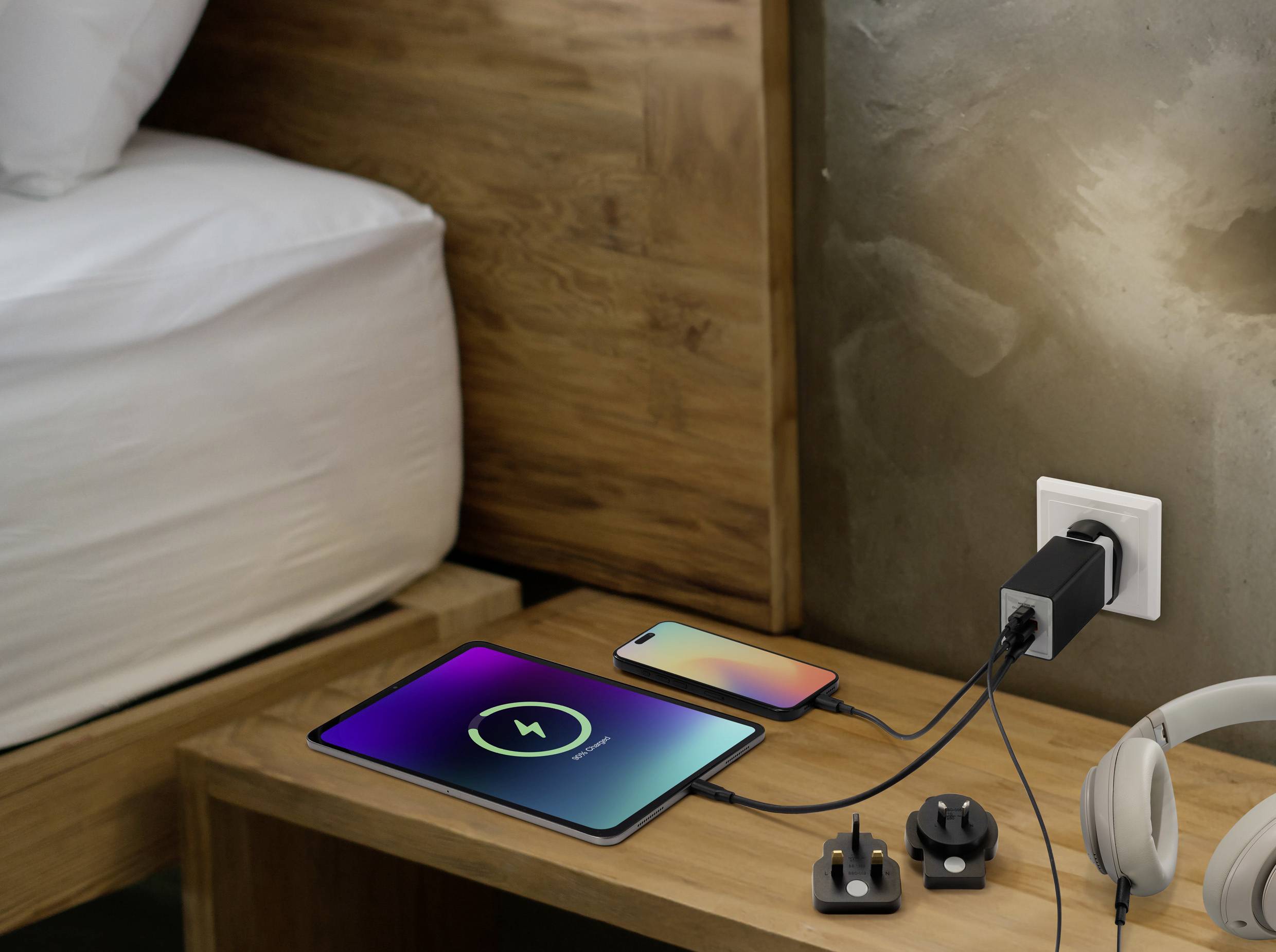 Tablet a smartphone se nabíjejí na dřevěném nočním stolku pomocí zástrčky s mezinárodními adaptéry; sluchátka jsou poblíž.
