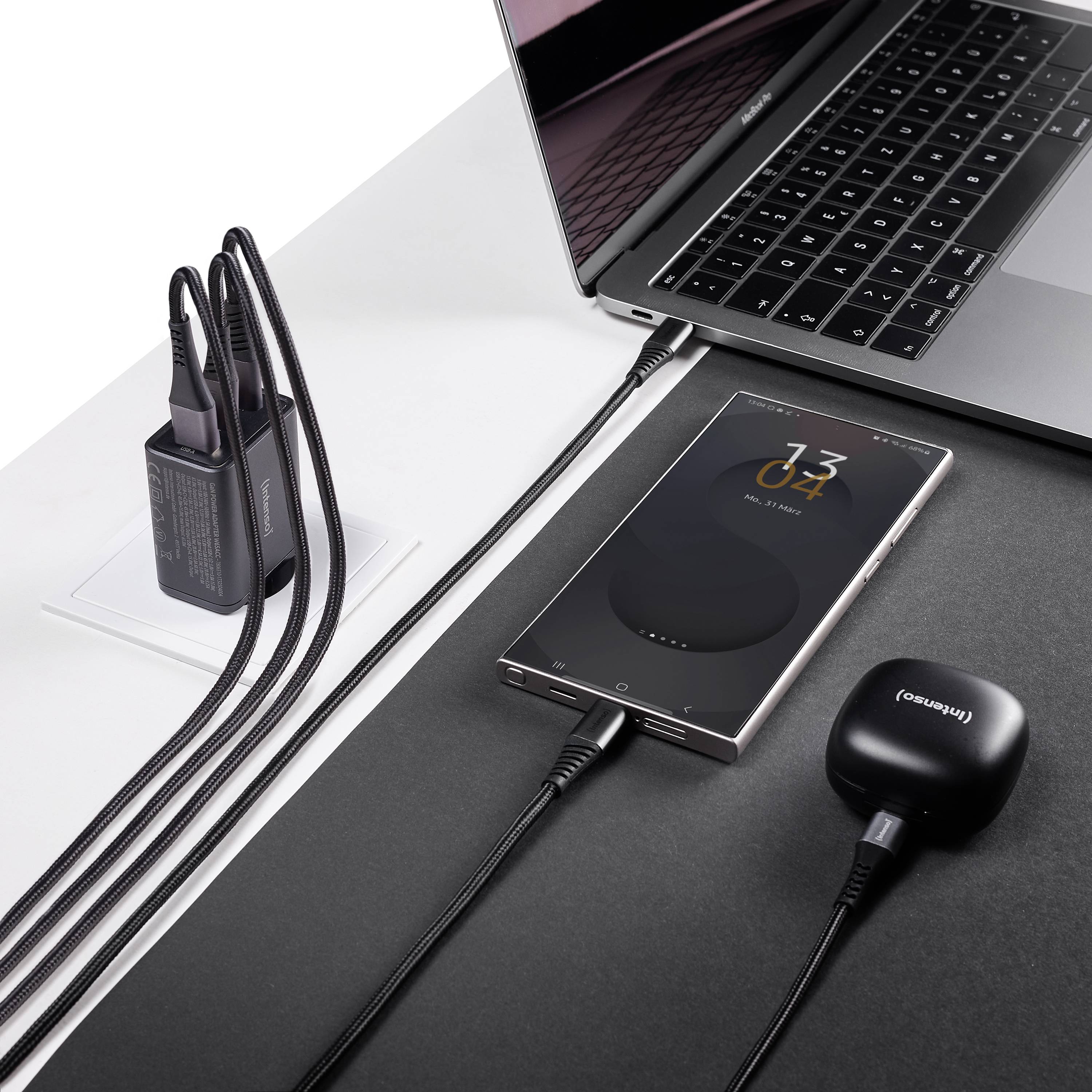 Smartphone bezdrátově nabíjející se na nabíjecí podložce vedle notebooku a zapojeného nabíječe.