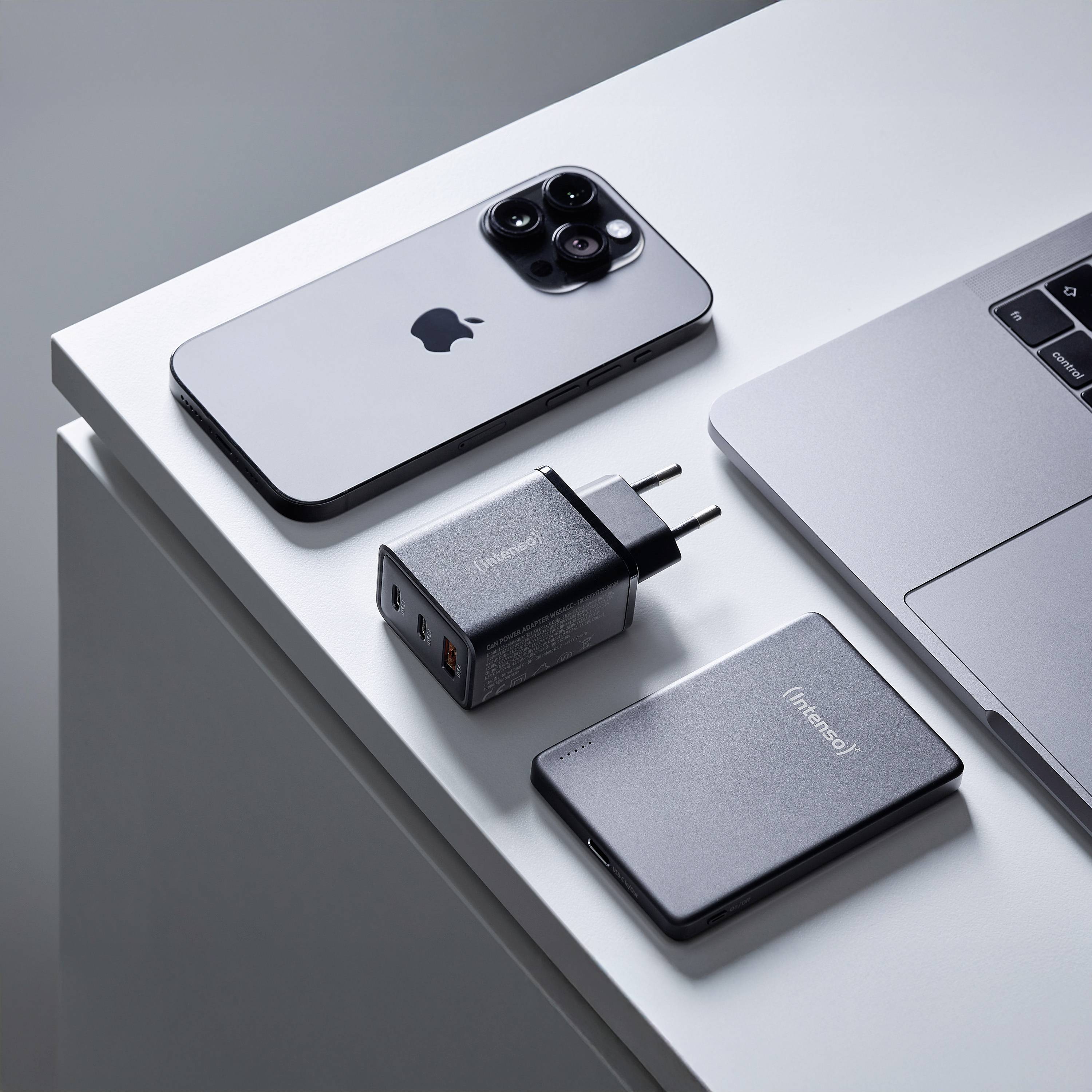 Smartphone, laptop, přenosná nabíječka a USB adaptér jsou uspořádány pečlivě na stole a ukazují minimalistické technologické vybavení.