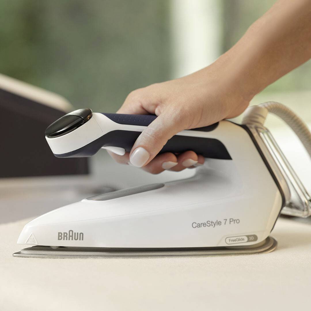 Ruka držící žehličku Braun CareStyle 7 Pro, umístěná na desce, demonstrující její ergonomický design.