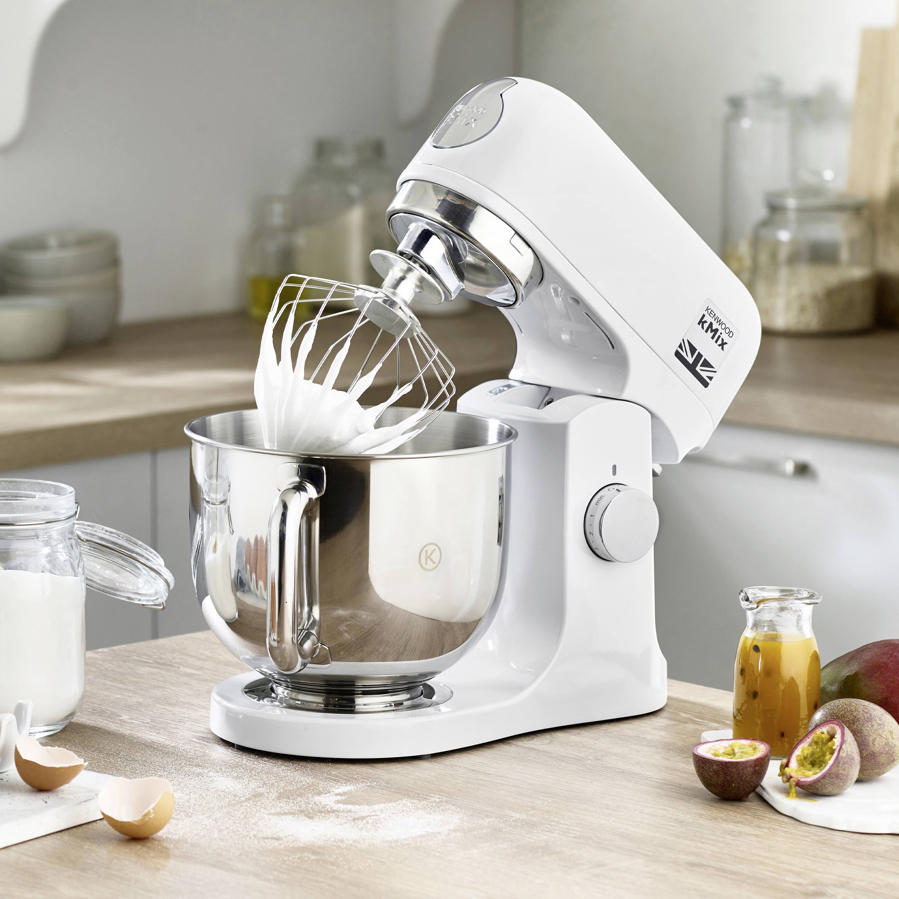 Bílý kuchyňský mixér se šlehací metlou šlehá smetanu v nerezové míse. Poblíž jsou ingredience a ovoce.