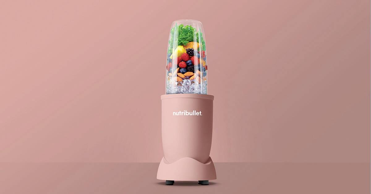Růžový mixér NutriBullet naplněný různými druhy ovoce a zeleniny, včetně bobulí a listové zeleniny, na pozadí v odpovídající růžové barvě.
