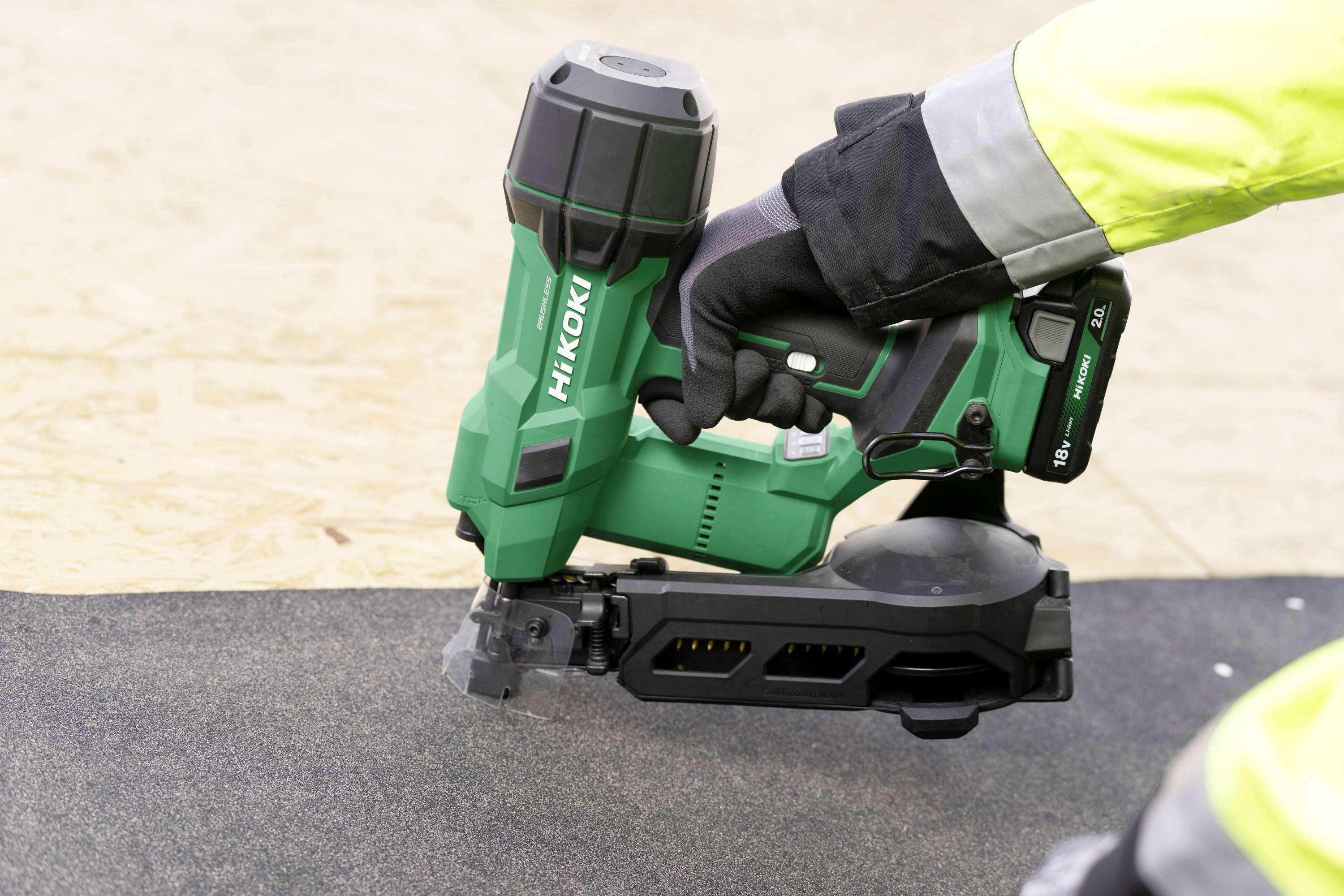 Osoba v reflexní vestě používá zelený hřebíkový přístroj Hitachi na černém povrchu, pravděpodobně střešním materiálu.