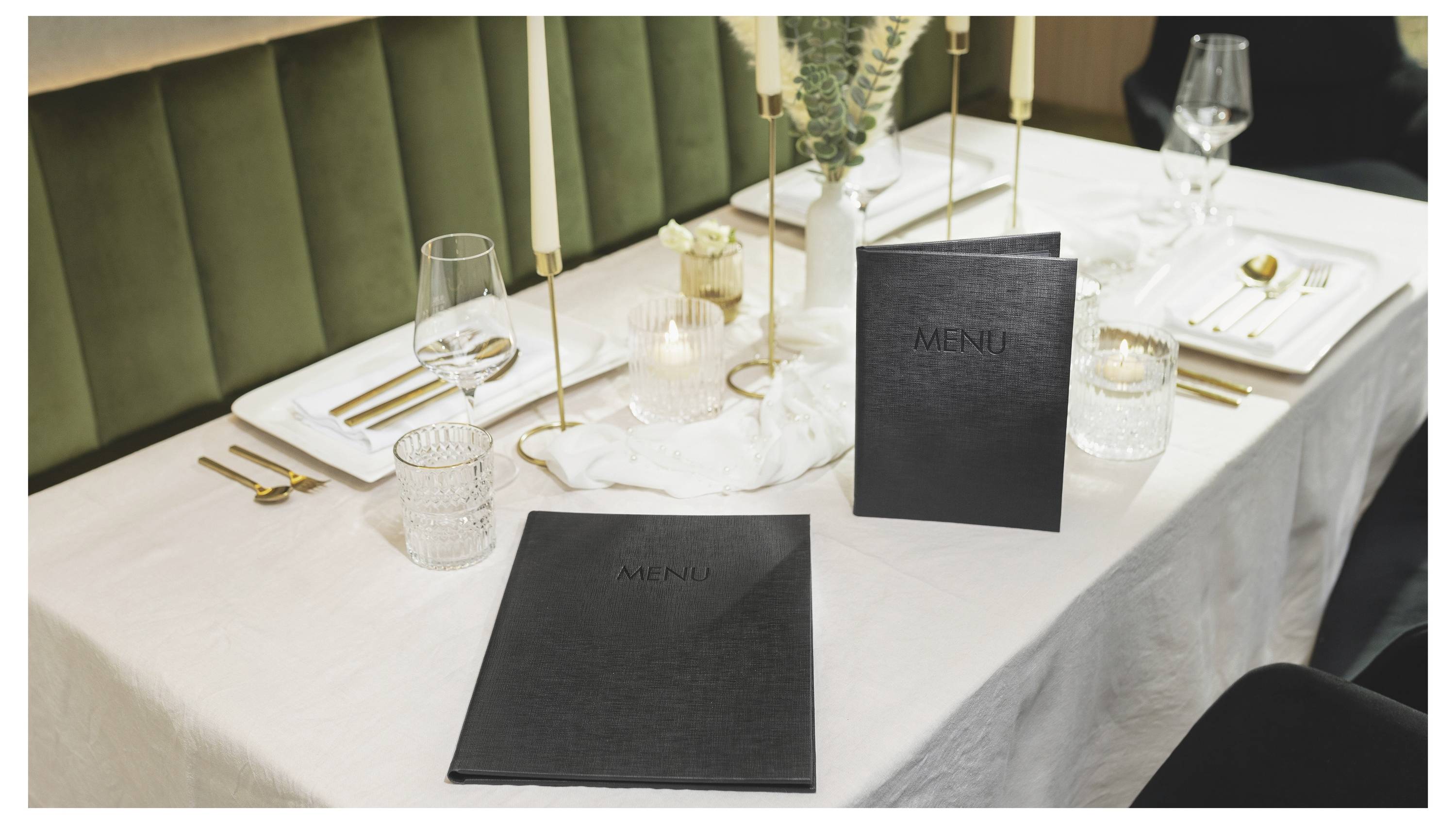 Elegantně upravený restaurační stůl s bílým prádlem, černými jídelními lístky, zlatými příbory, sklenicemi, svíčkami a květinovou dekorací ve středu.